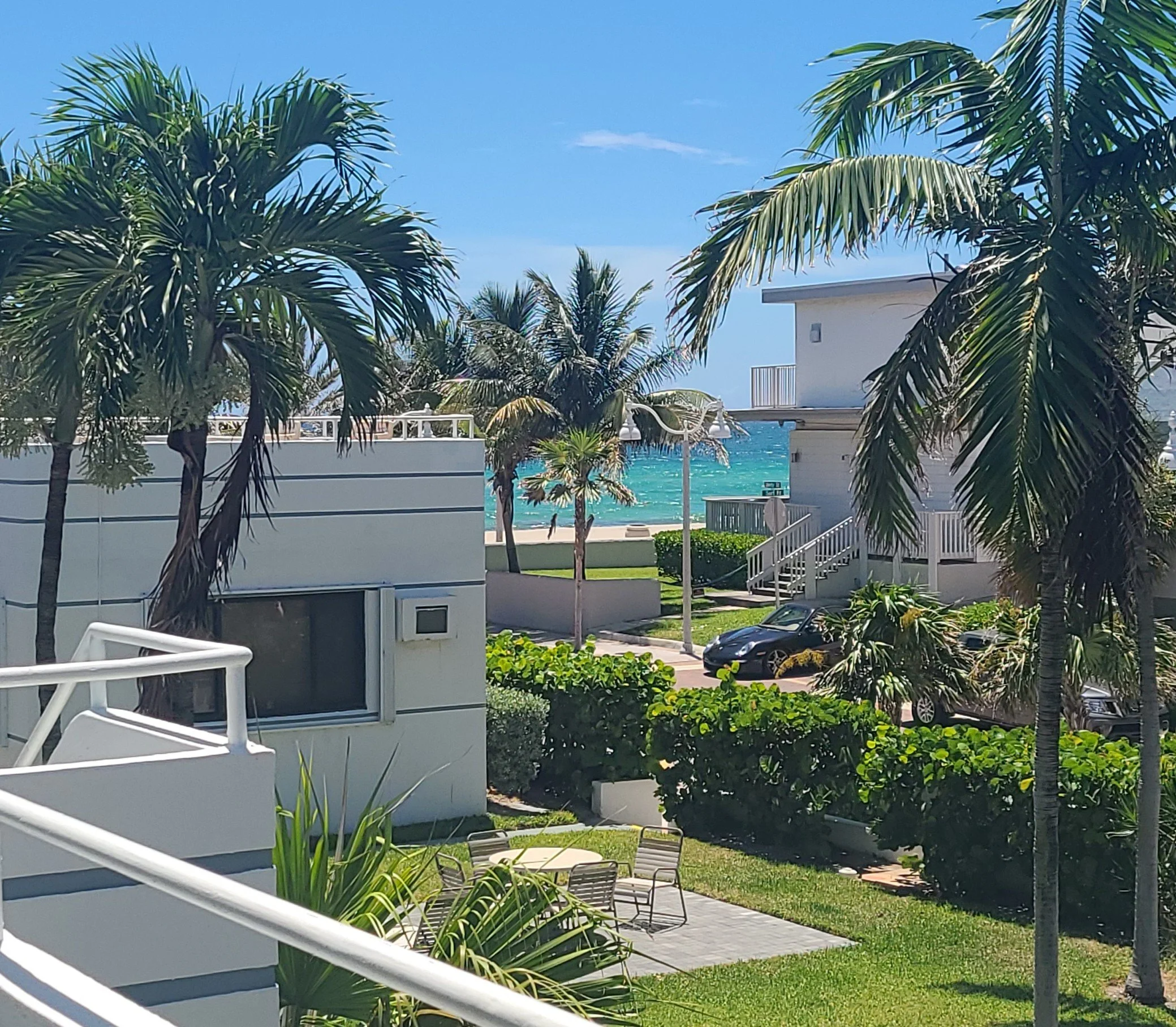View of a sunny coastal area with white buildings, palm trees, a sidewalk, parked cars, a grassy lawn with patio furniture, and the ocean in the background.