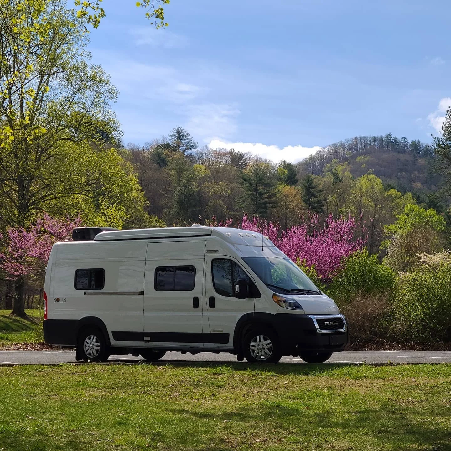 A white RV parked on a grassy area along a road with lush green trees and pink blooming trees in the background, hills and a partly cloudy blue sky in the distance.