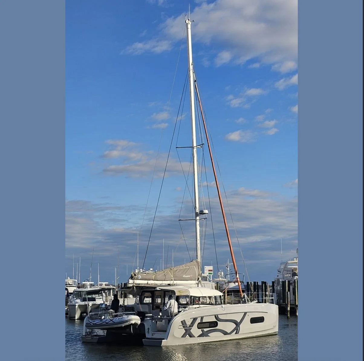 A sailboat docked at a marina with multiple other boats, under a blue sky with scattered clouds.
