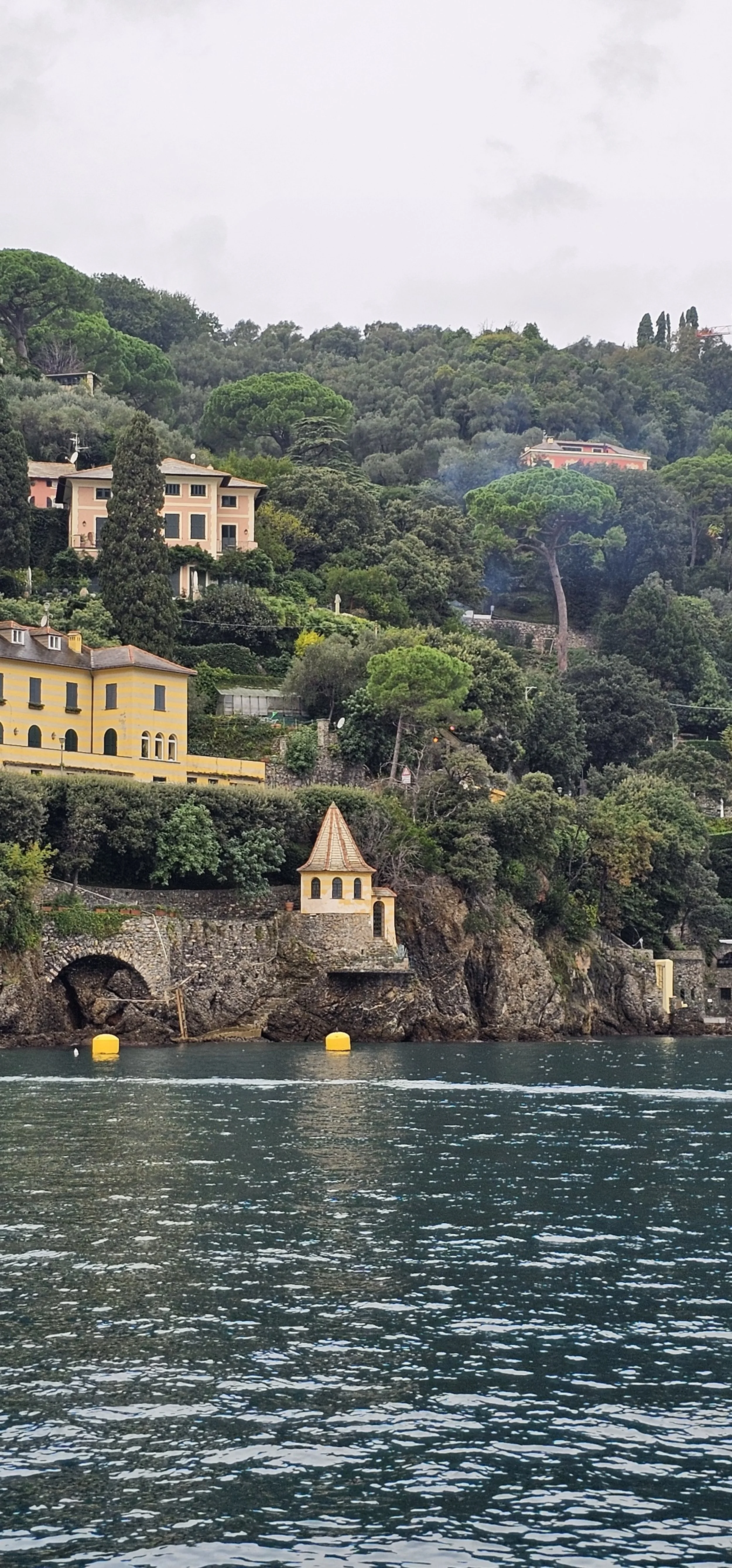 A coastal hillside with various colorful houses and lush green trees, a small chapel near the water, and buoys floating on the water's surface.