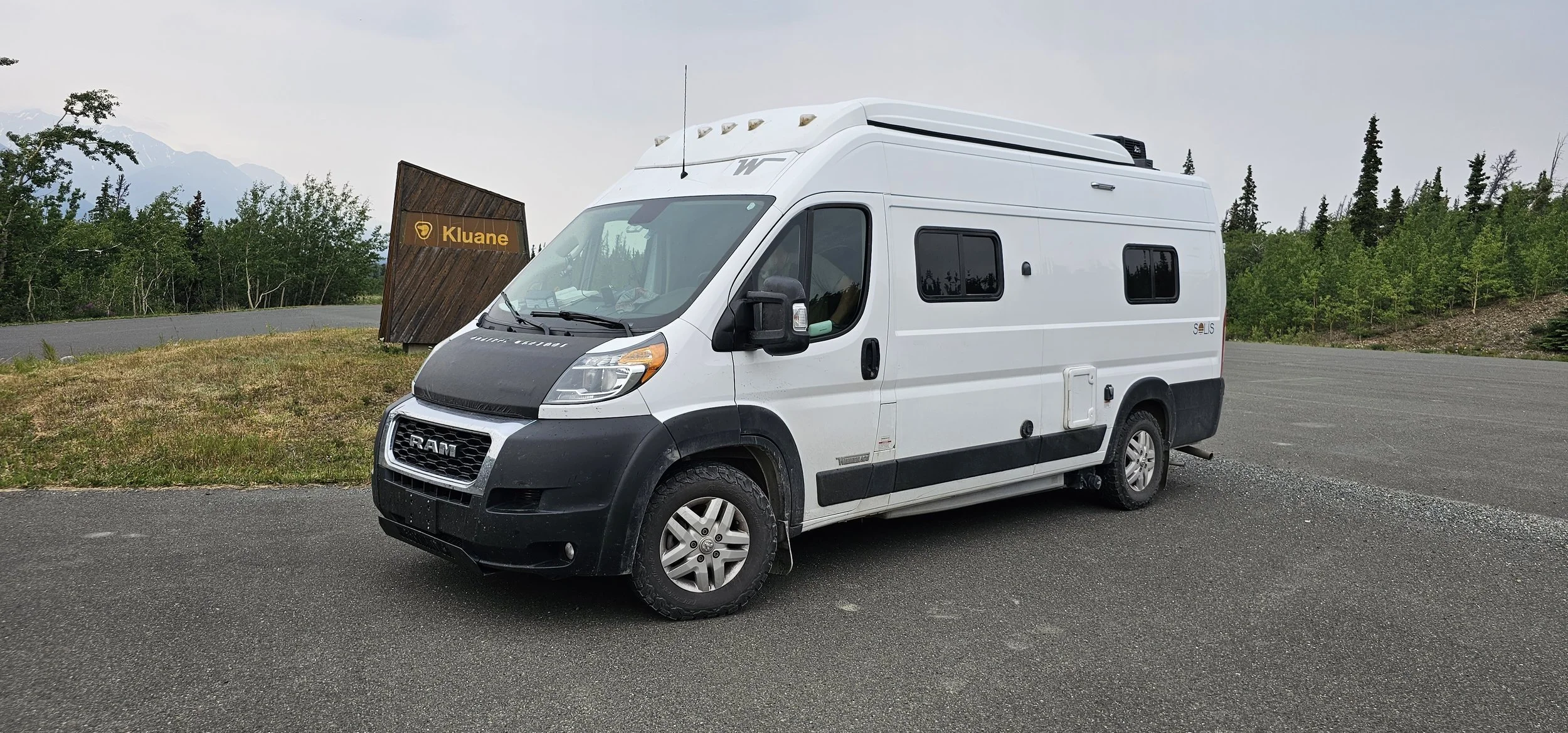 A white Ram camper van parked on an asphalt area near a small grassy patch with trees and mountains in the background, and a sign that reads 'Kluane'.