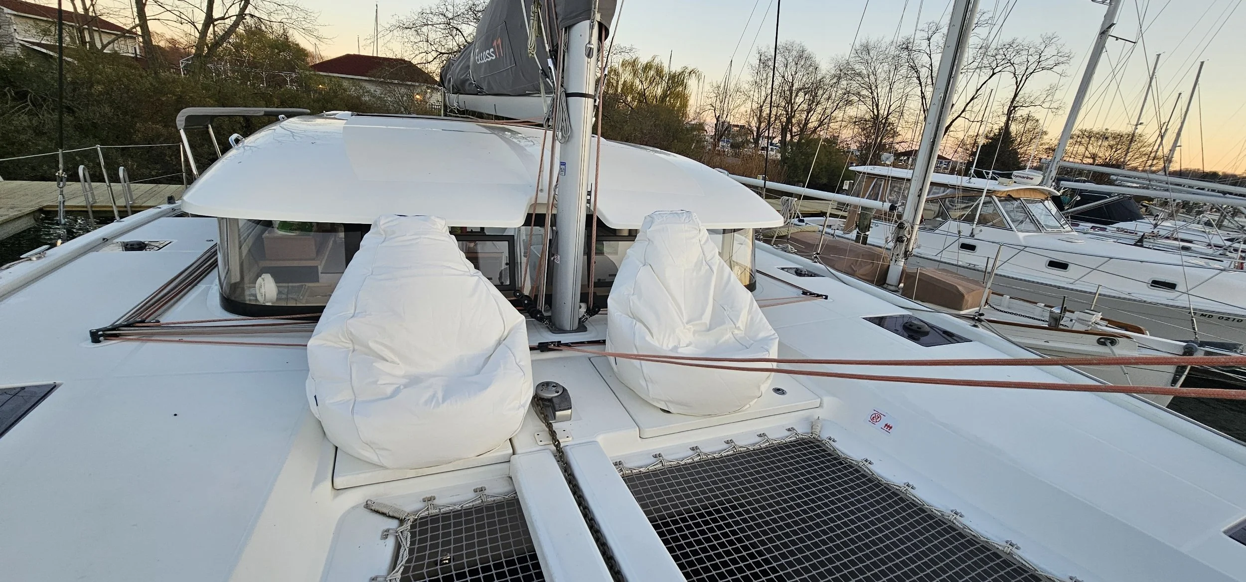 View of a white sailing catamaran docked at a marina during sunset with other boats nearby and leafless trees in the background.