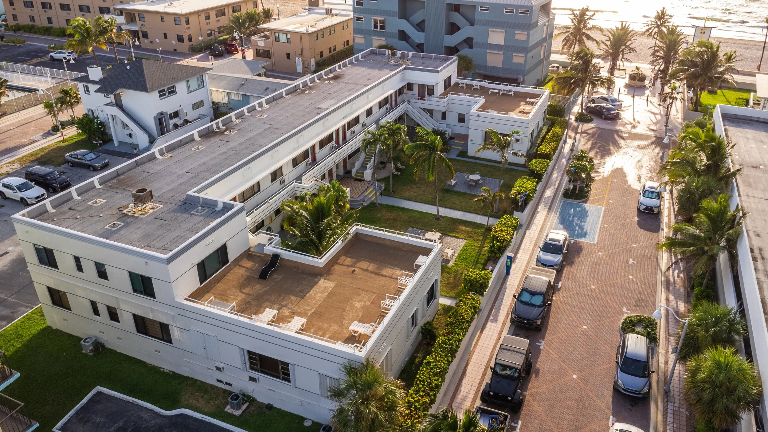 An aerial view of a beachside apartment complex with courtyard, surrounded by parked cars, palm trees, and neighboring buildings, near a sandy beach and ocean.