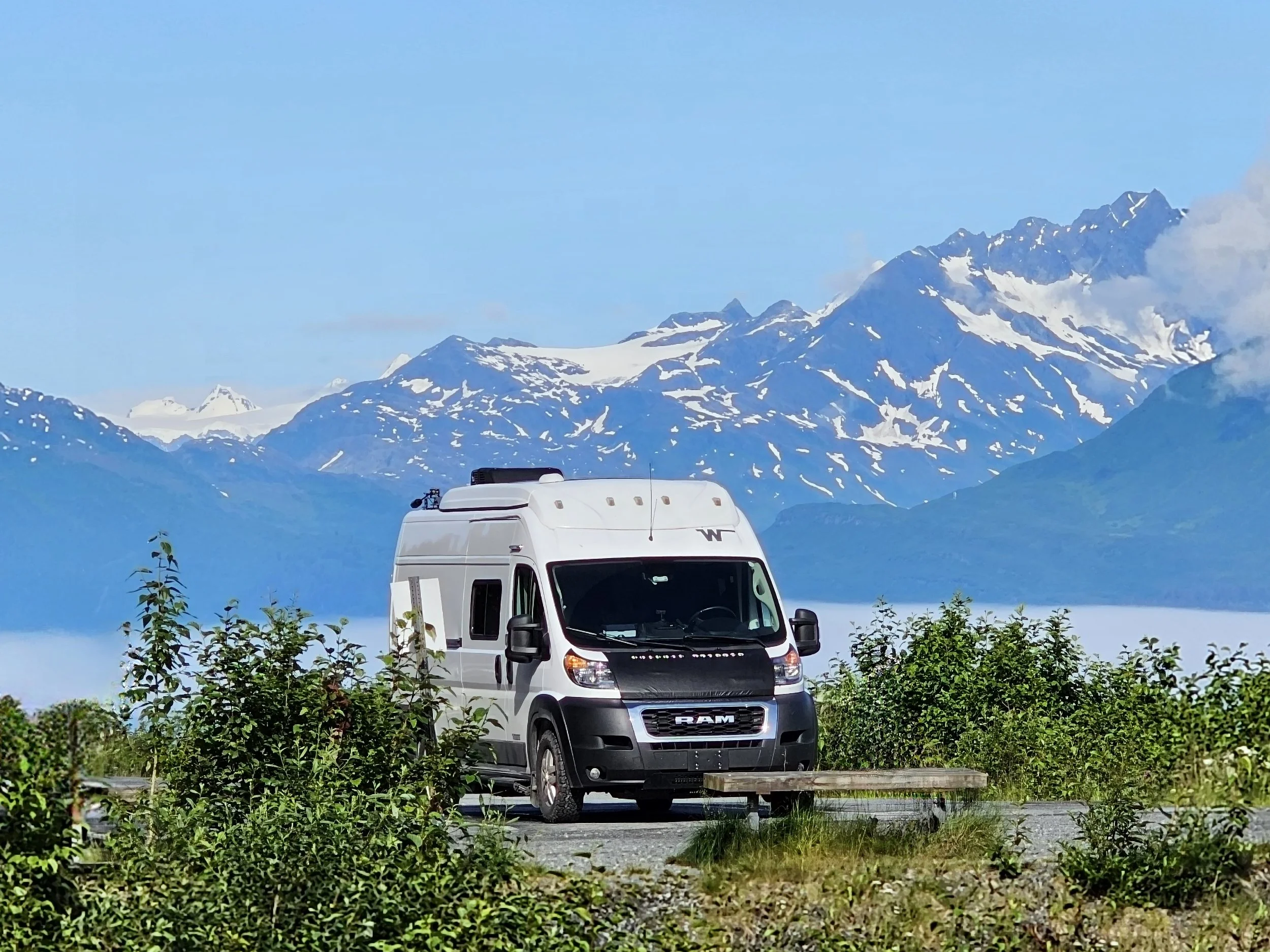 A white camper van parked in a scenic mountainous area with snow-capped peaks in the background, greenery in the foreground, and a clear blue sky.