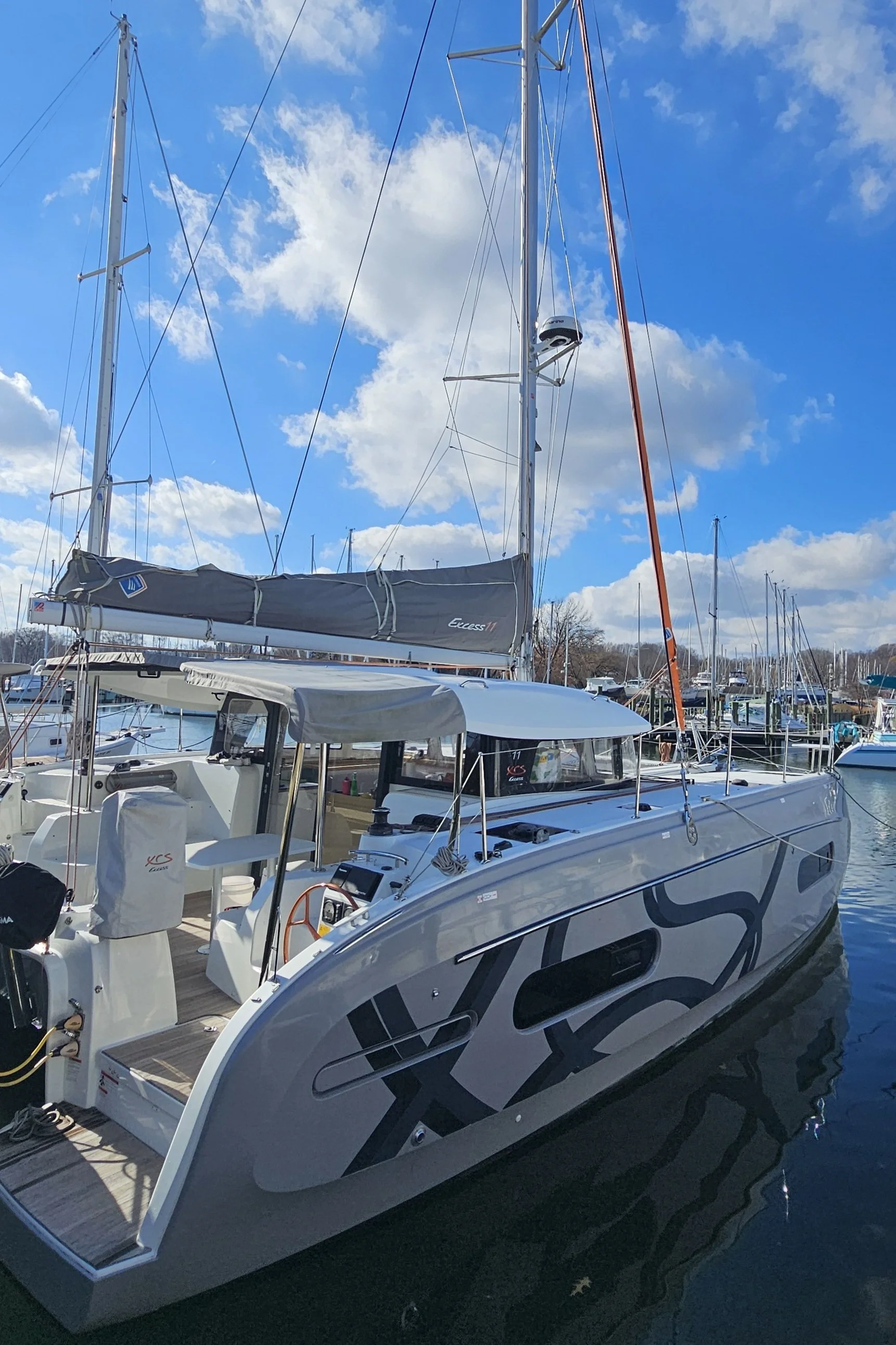 A modern sailboat docked in a marina with other boats, under a partly cloudy blue sky.