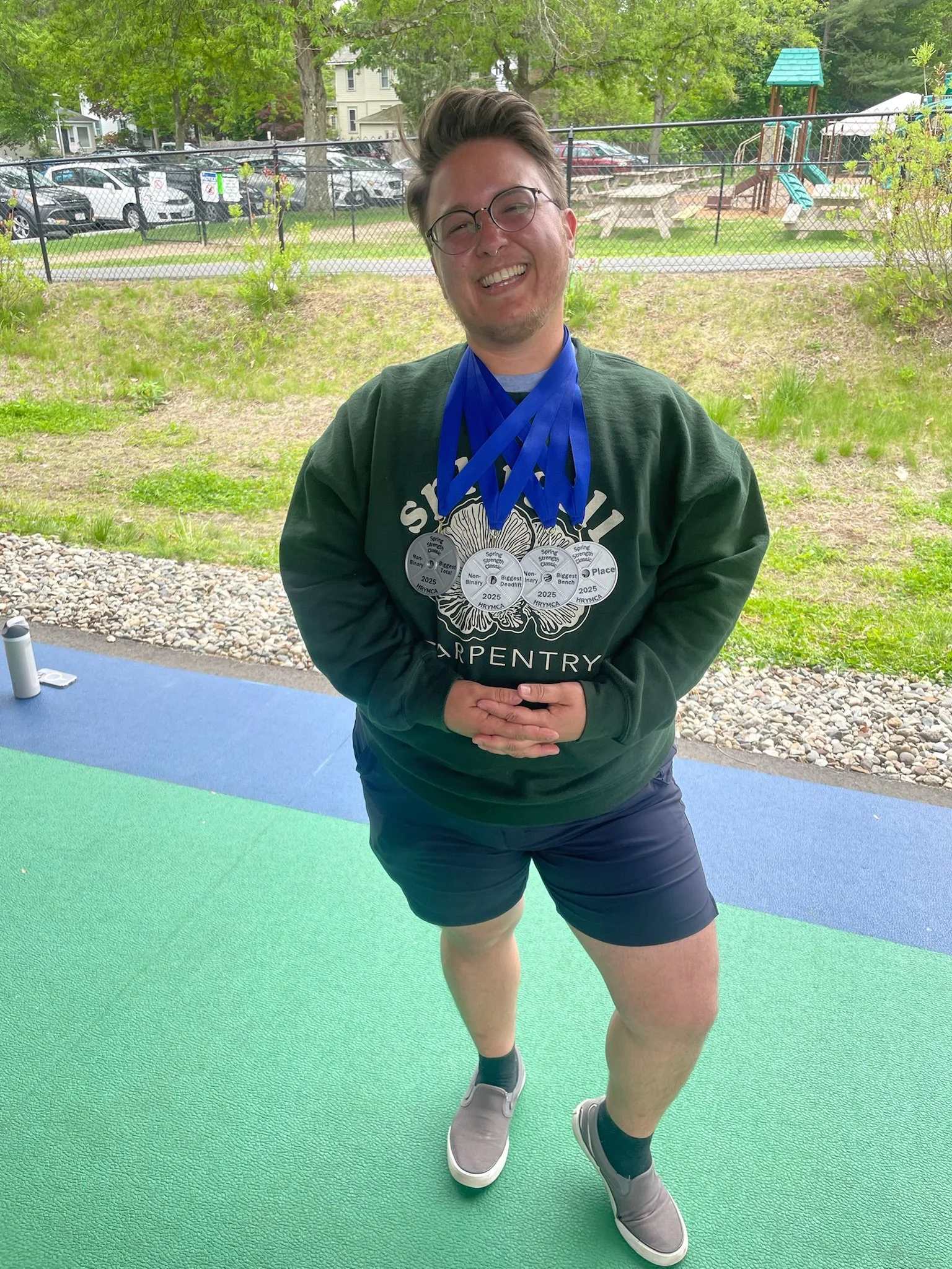 A young man standing on a green and blue outdoor surface, wearing shorts, a sweatshirt, and sneakers, smiling, with medals hanging around his neck, outdoors in a park or backyard setting.