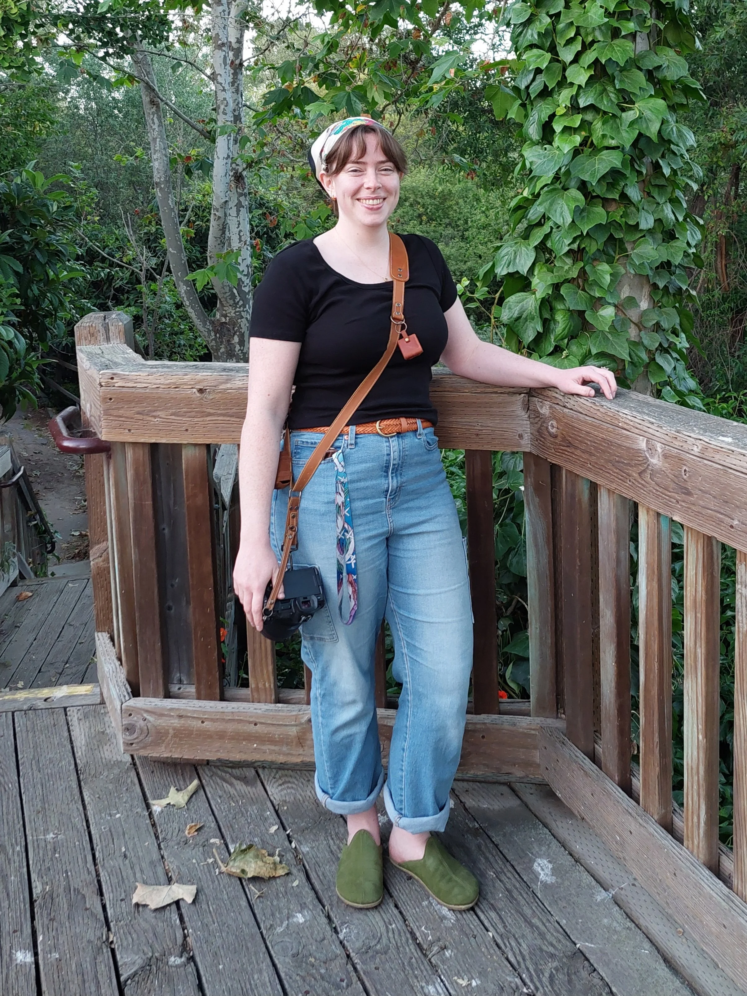 Young woman with short brown hair, wearing a black t-shirt, light blue rolled-up jeans, green slippers, and a patterned headscarf, standing on a wooden deck with a forest background, smiling and leaning on the railing.