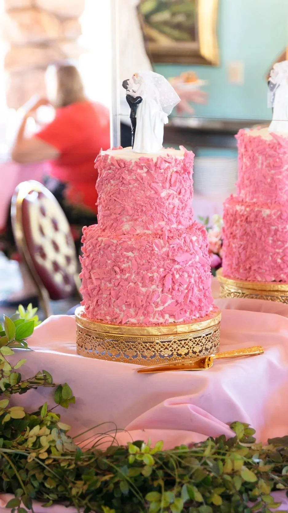 Pink three-tiered wedding cake from the Madonna Inn with shredded pink decorations, topped with a bride and groom figurine, placed on a pink cloth with greenery and a gold cake server in front.