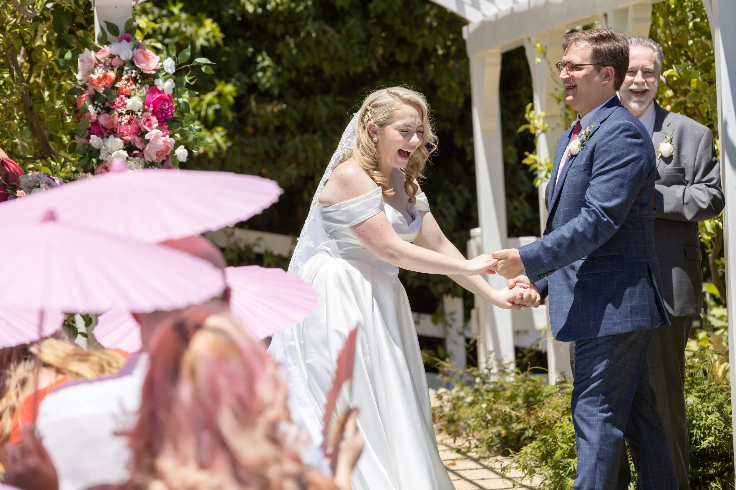 A bride and groom holding hands and smiling during their outdoor wedding ceremony, with a joyful officiant in the background. Guests are visible in the foreground under pink umbrellas.