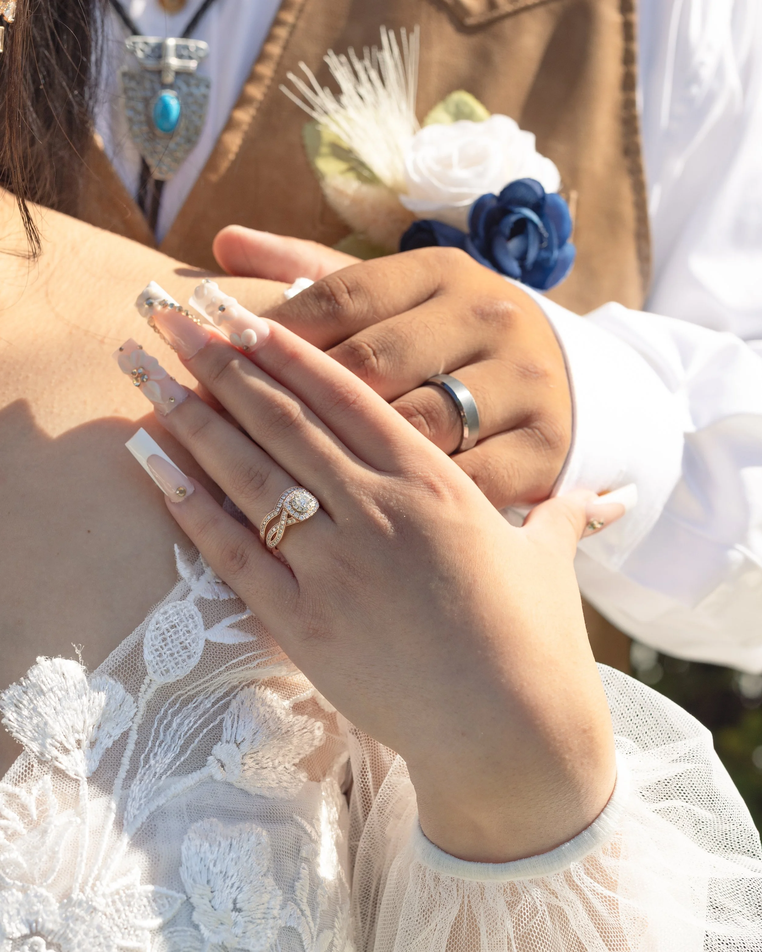 Close-up of a couple's hands with wedding rings, hands resting on a woman's shoulder, wearing wedding attire, with a boutonniere pinned to the man's shirt.