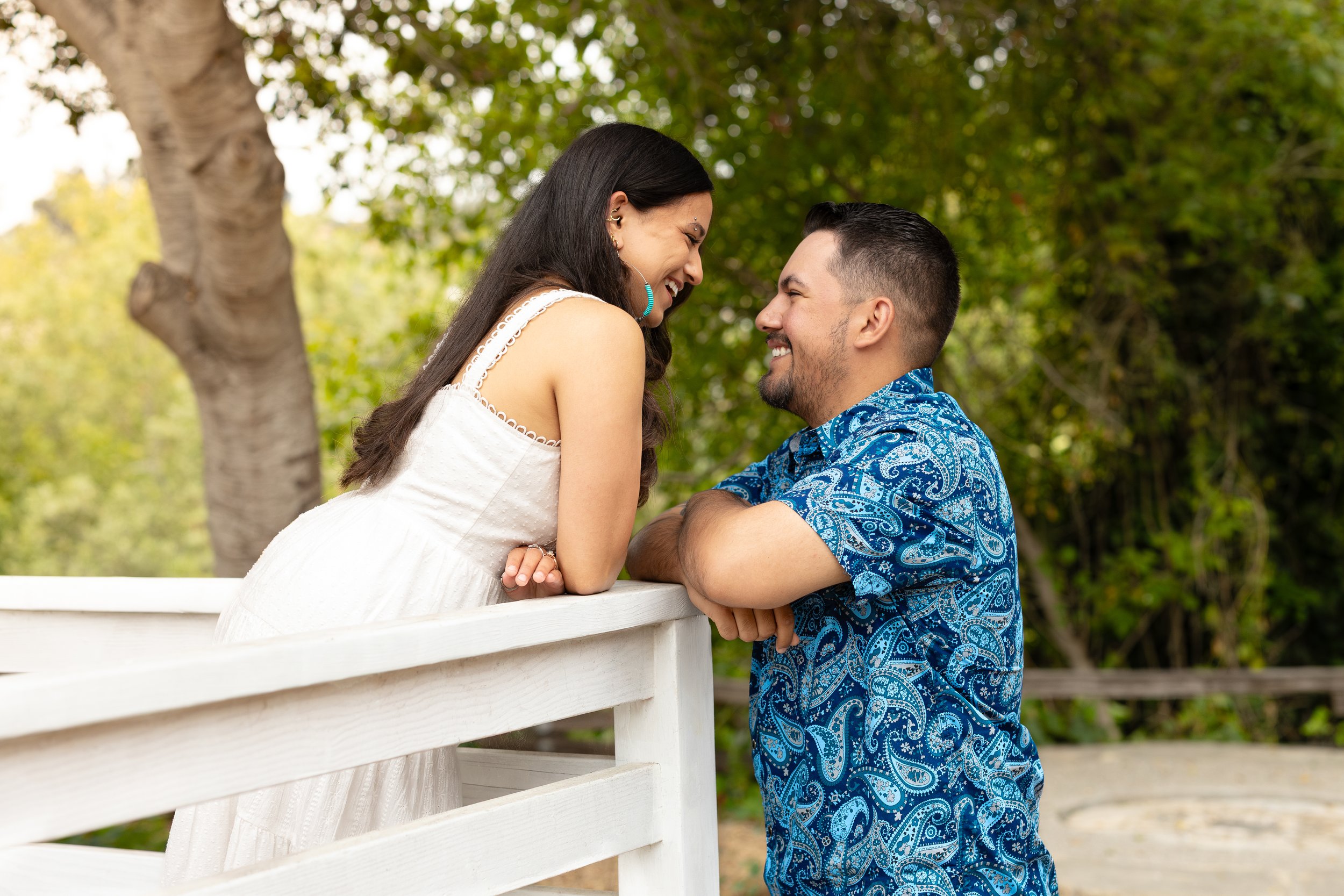 A couple leaning on a white wooden railing outdoors, smiling and looking into each other's eyes, with green trees in the background.