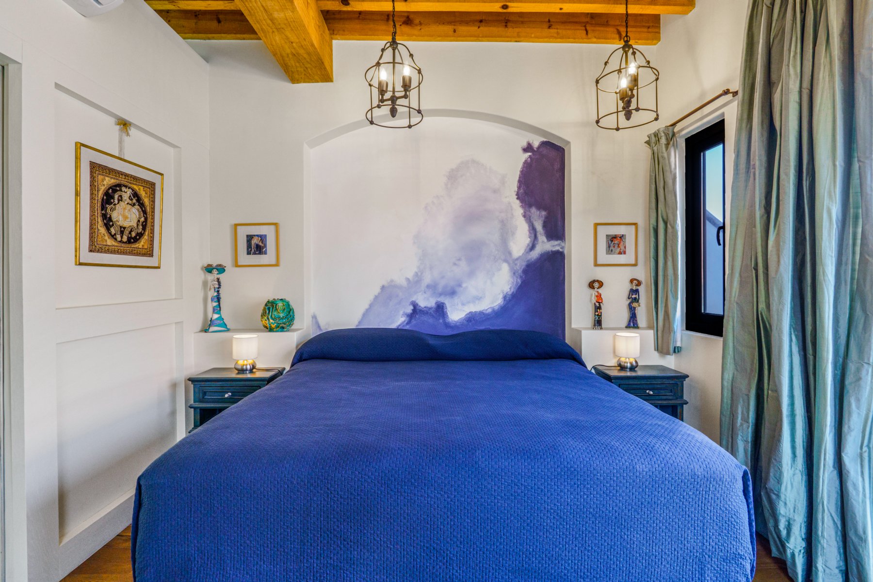 A bedroom with white walls, wooden beams, and a large bed with a blue bedspread. There are two small nightstands with lamps, colorful decorative objects, and framed artwork. A window with curtains is on the right side, and there's a mountain-themed m