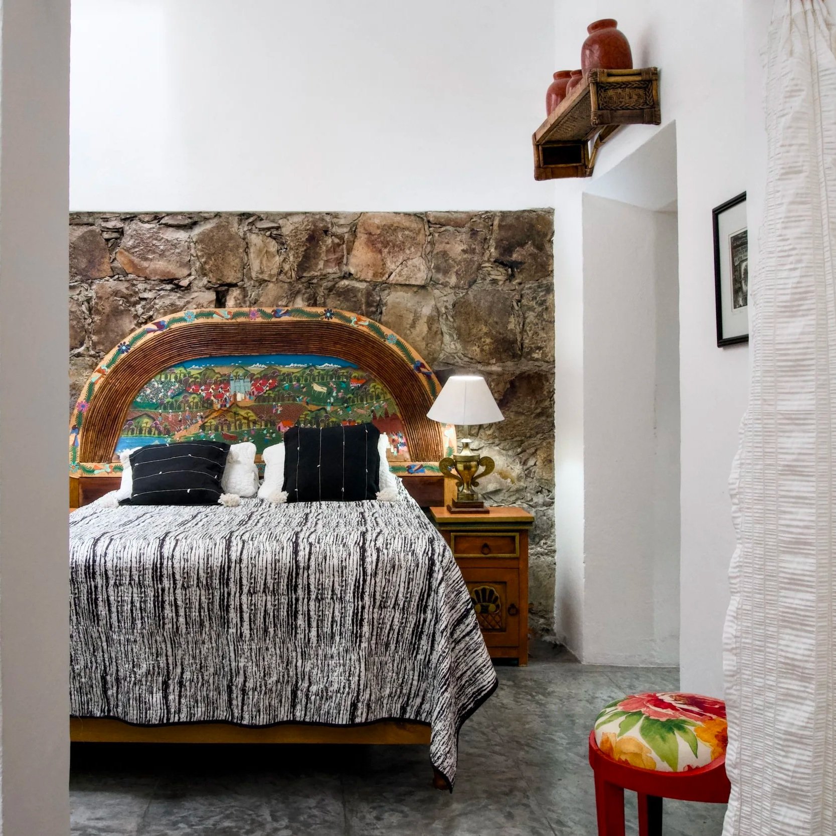 A bedroom with a bed featuring a colorful, intricate headboard, black and white striped bedding, and three pillows. There is a wooden nightstand with a lamp beside the bed, a stone wall behind the bed, and white walls with a small decorative shelf an