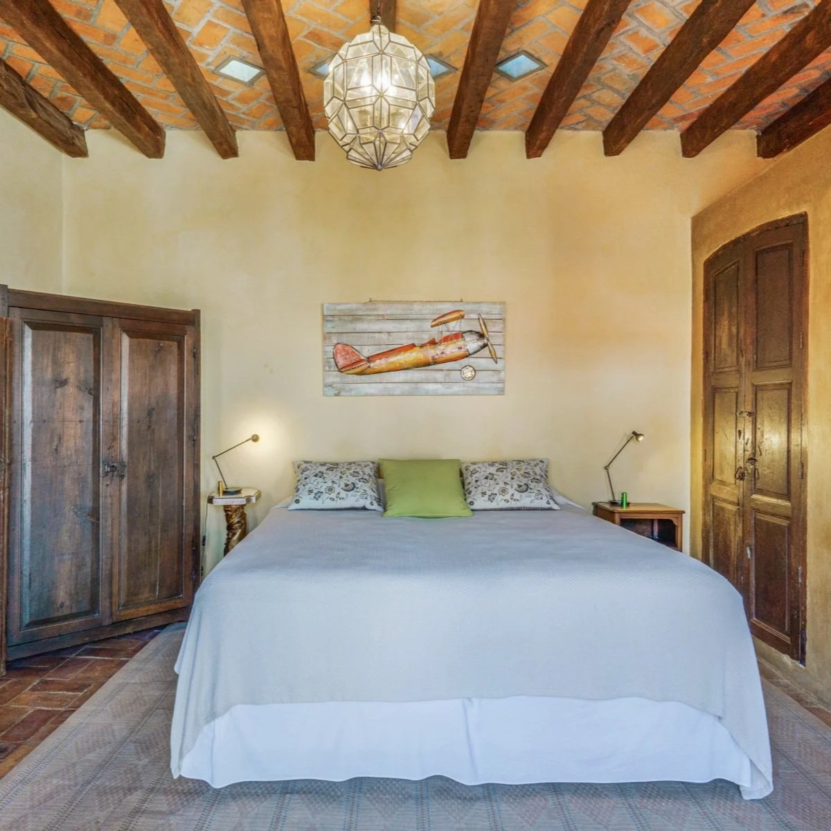 A cozy bedroom with a large bed covered in white bedding with three pillows, two with floral patterns and one green. Above the bed hangs a wooden art piece of a vintage airplane. On each side of the bed are wooden nightstands with adjustable reading 