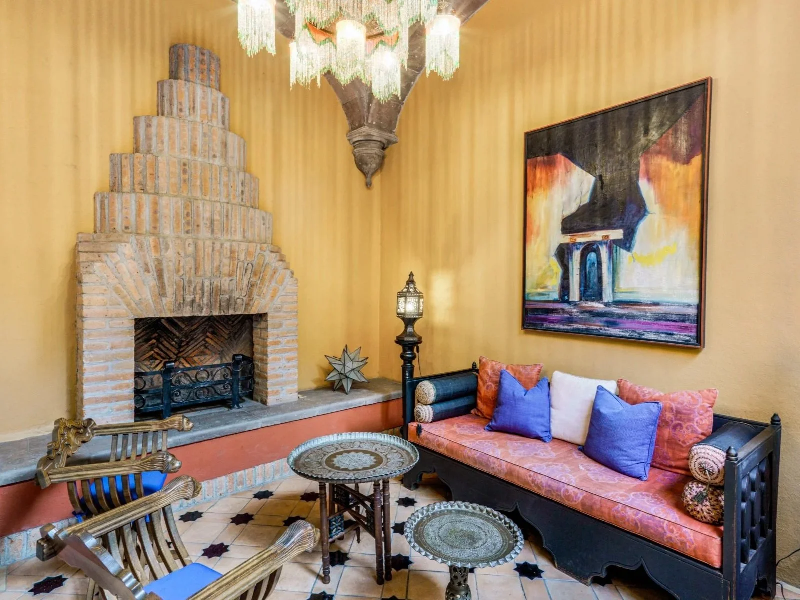 A living room with a brick fireplace, a colorful painting on the wall, a vintage black sofa with red and white cushions, two small ornate tables, and a chandelier hanging from the ceiling.