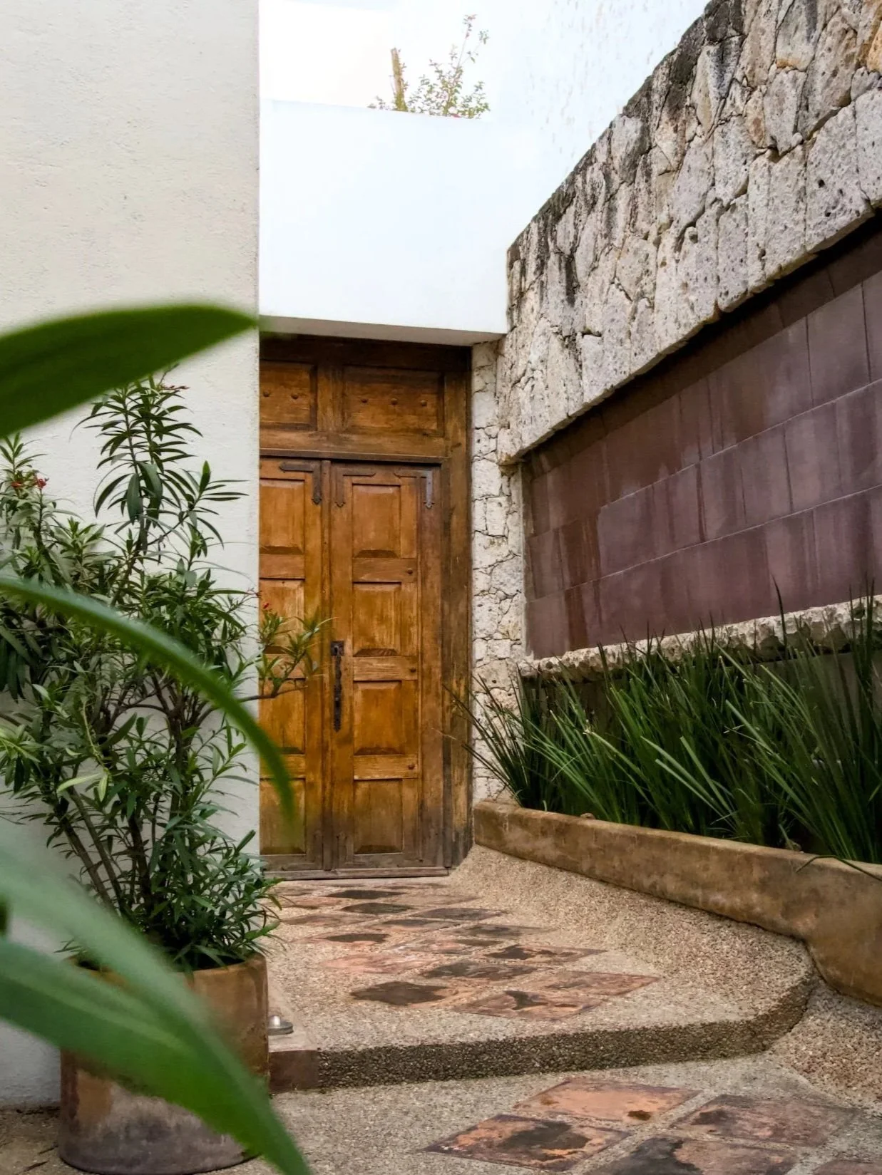 A wooden door with a stone and tile wall on one side, potted plants, and a curved stone pathway in an outdoor courtyard.
