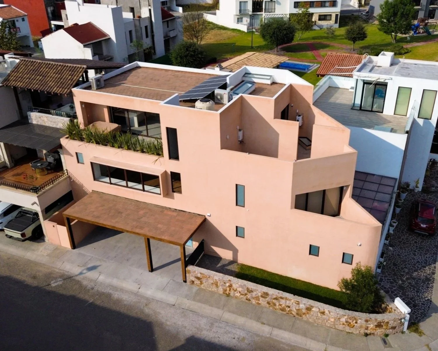 A modern pink multi-story house with a flat roof, large windows, and outdoor terrace areas, situated in a neighborhood with other residential buildings and greenery.