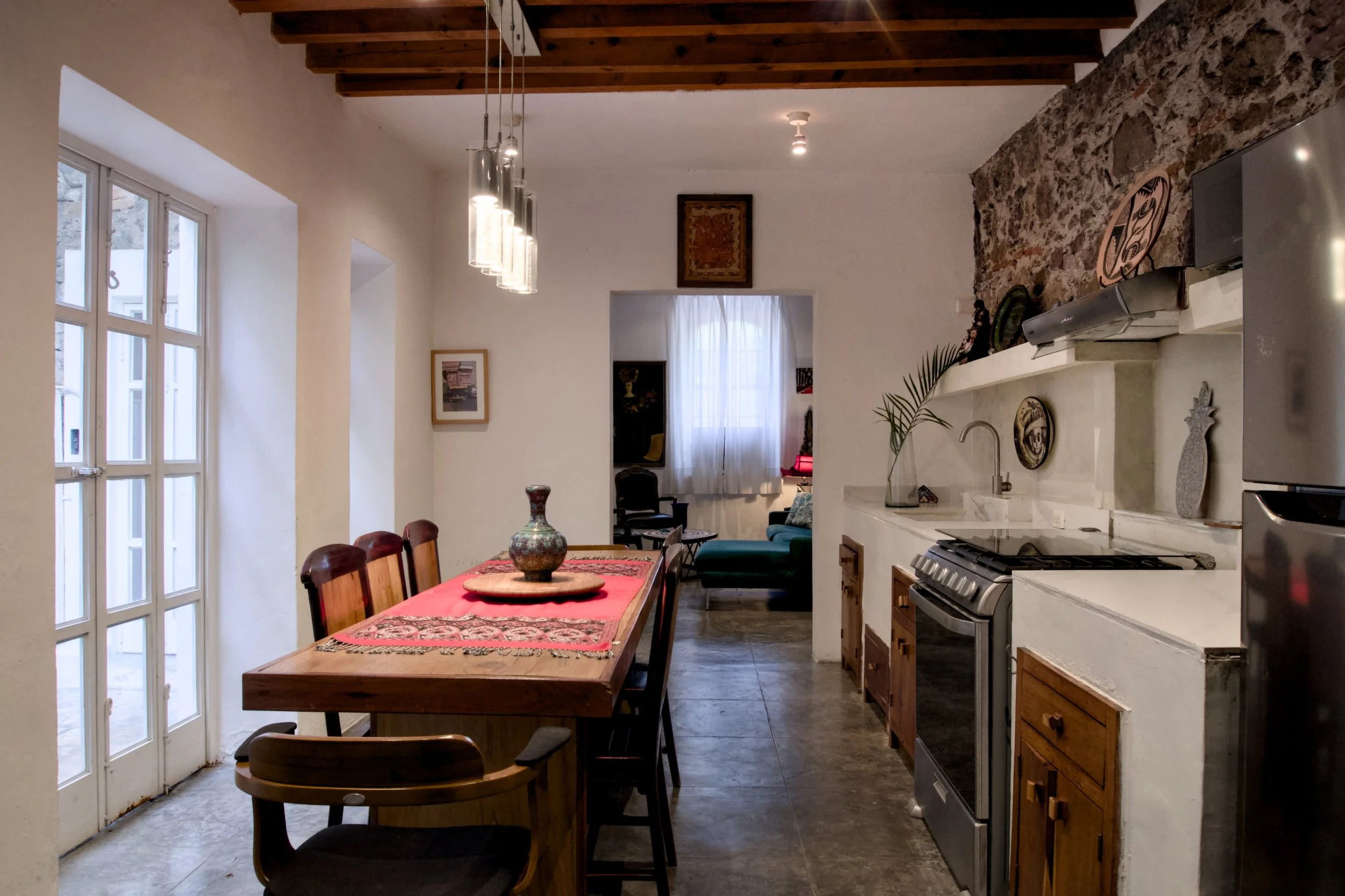 Interior view of a cozy kitchen and dining area with a wooden dining table, red table runner, decorative vase, white walls, a window, and a partially visible living room with green sofa and furniture.