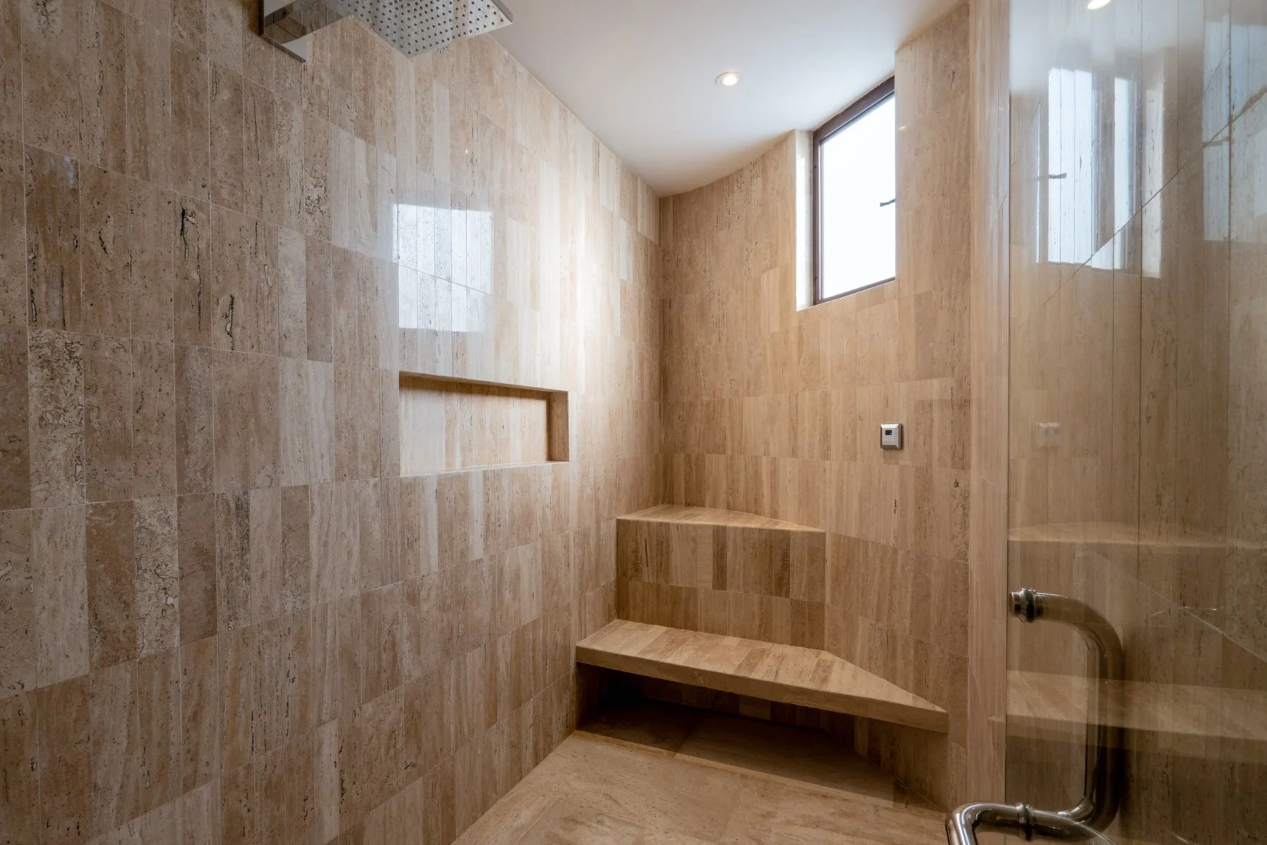 An unfinished shower area with beige tile walls, built-in corner seating, a small built-in niche, a window, and exposed plumbing.