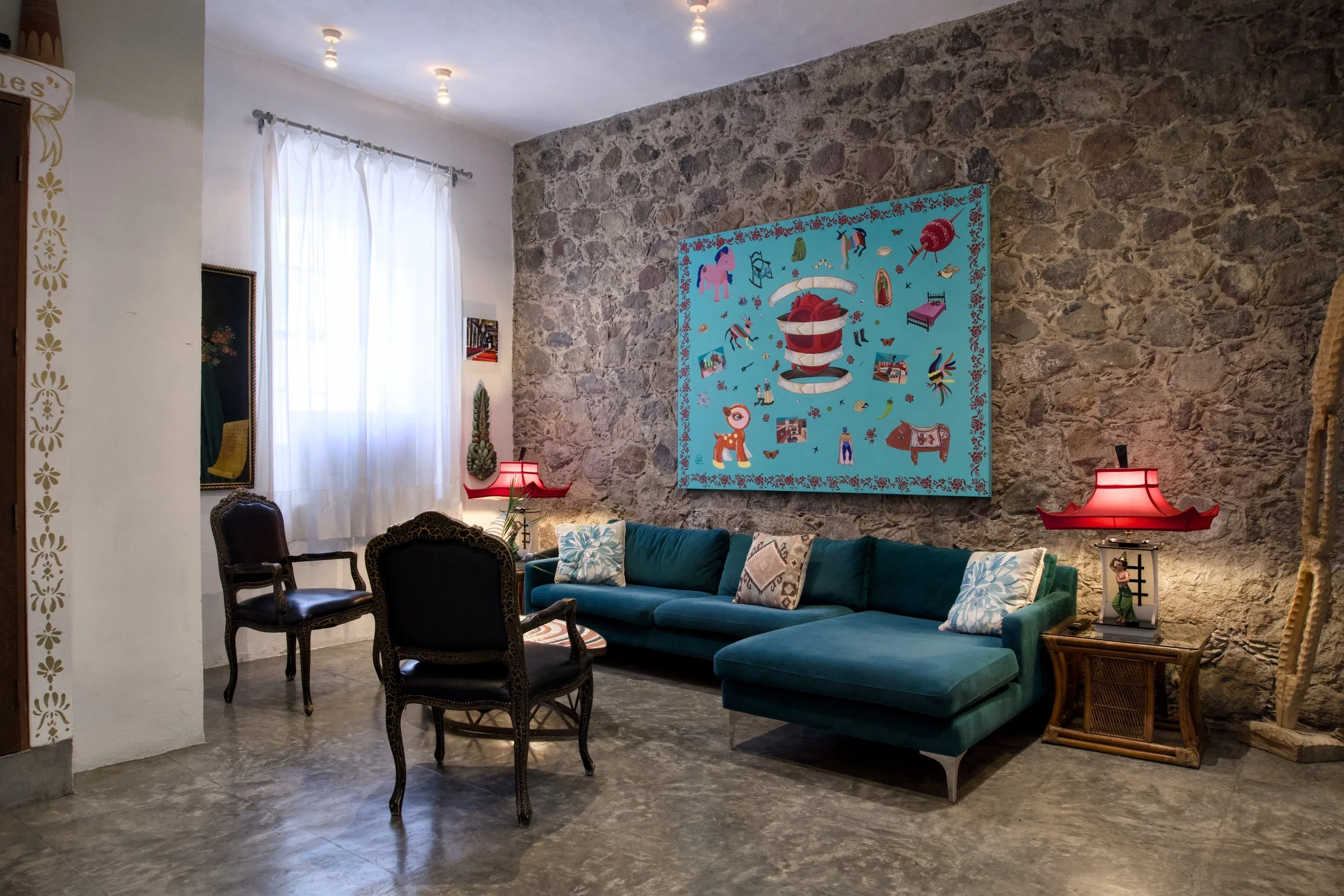 A cozy living room with a stone accent wall, a large teal sectional sofa with patterned pillows, a wooden side table with a red lamp, and eclectic decor including colorful artwork and vintage-style chairs.