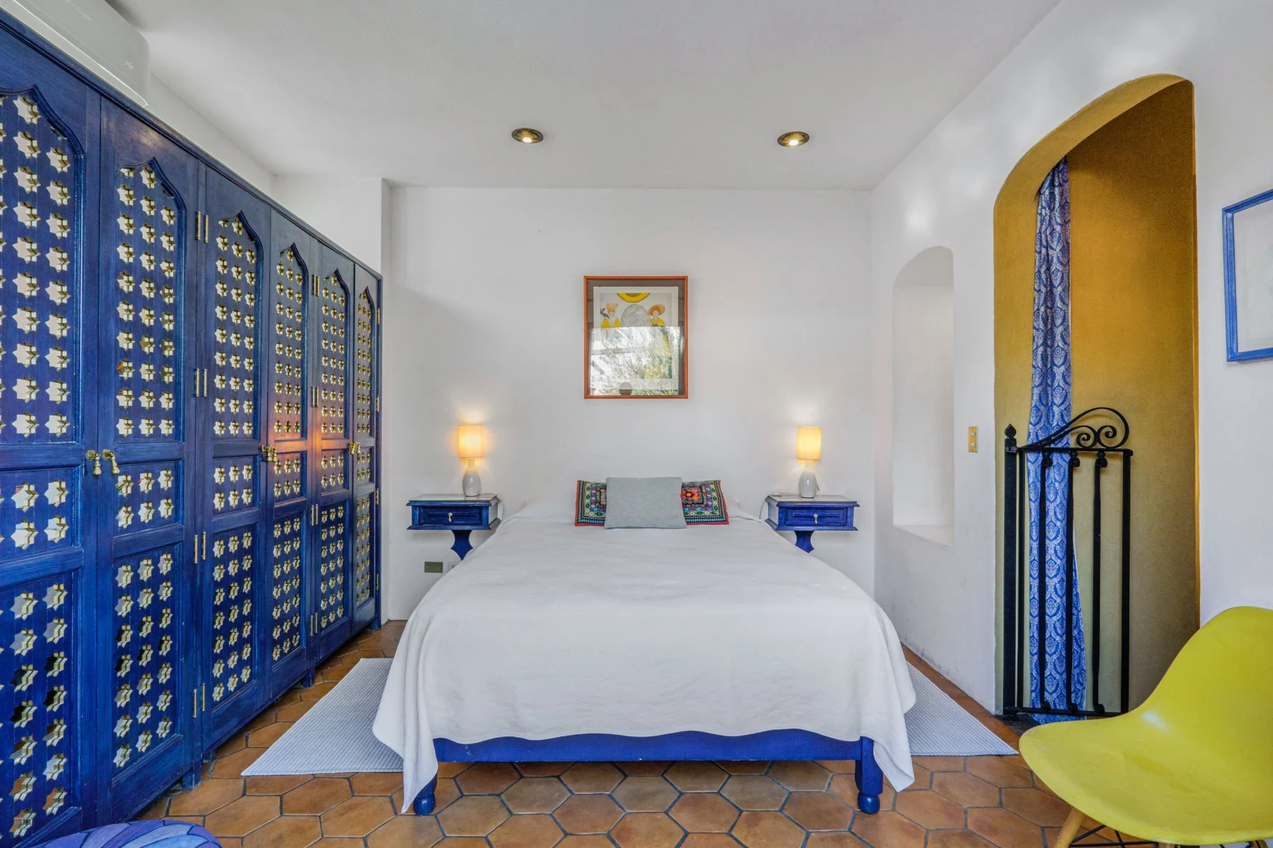 Bedroom with white walls and ceiling, a bed with white bedding, blue nightstands, yellow lamps, colorful pillows, a large blue wardrobe, and yellow accents in the decor.