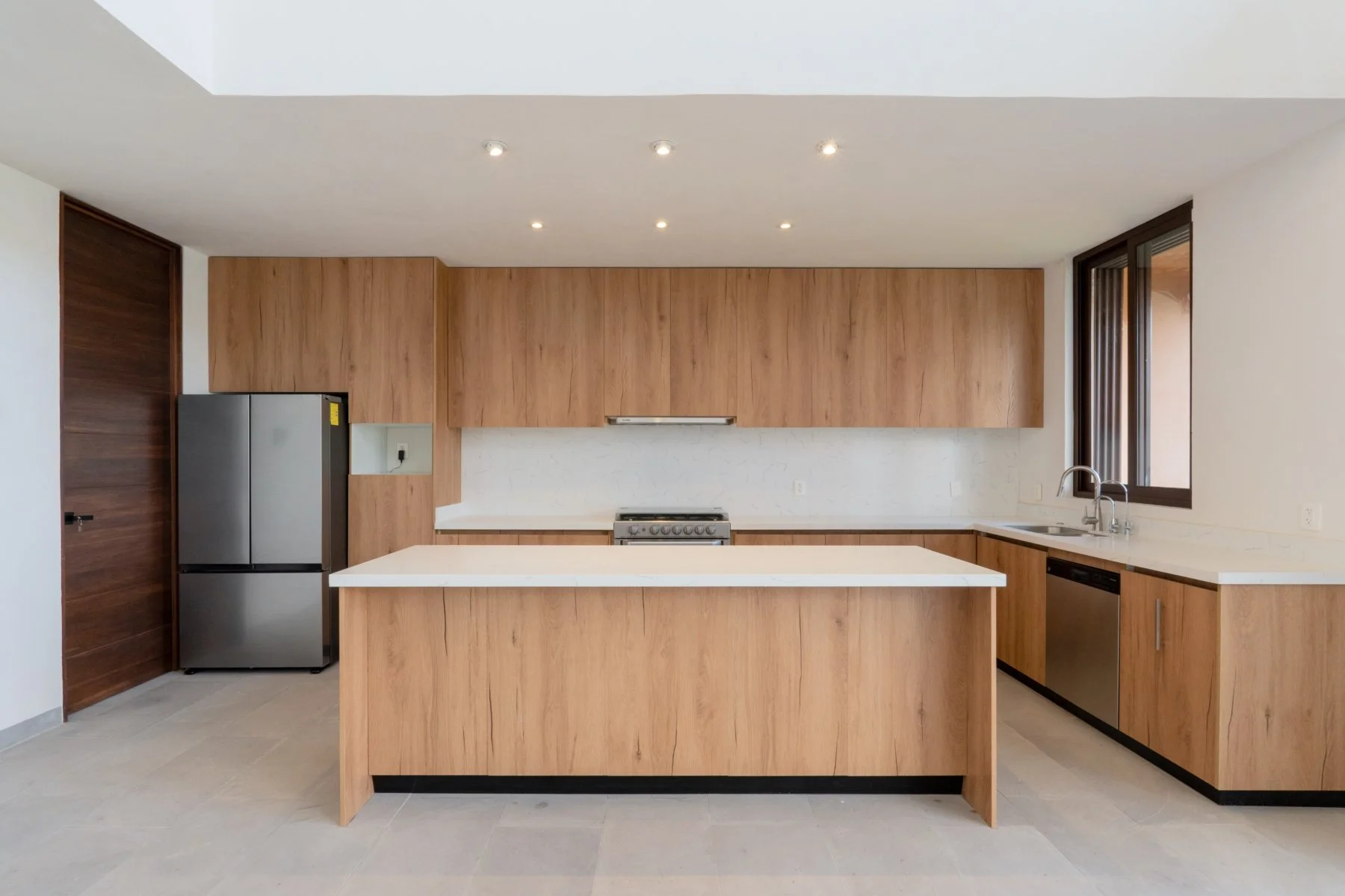 Modern kitchen with wooden cabinets, white countertops, stainless steel refrigerator, built-in oven, dishwasher, double sink, and large window.