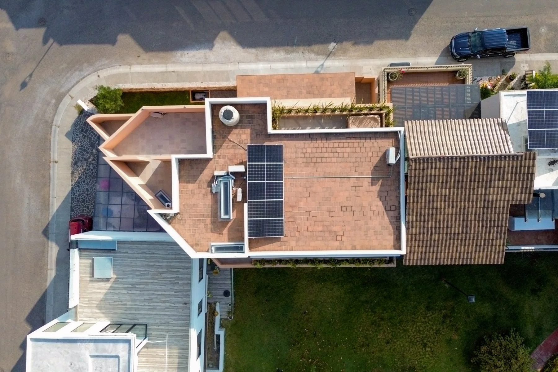 Bird's-eye view of residential rooftops with solar panels, brick and tile roofs, and nearby parked cars.