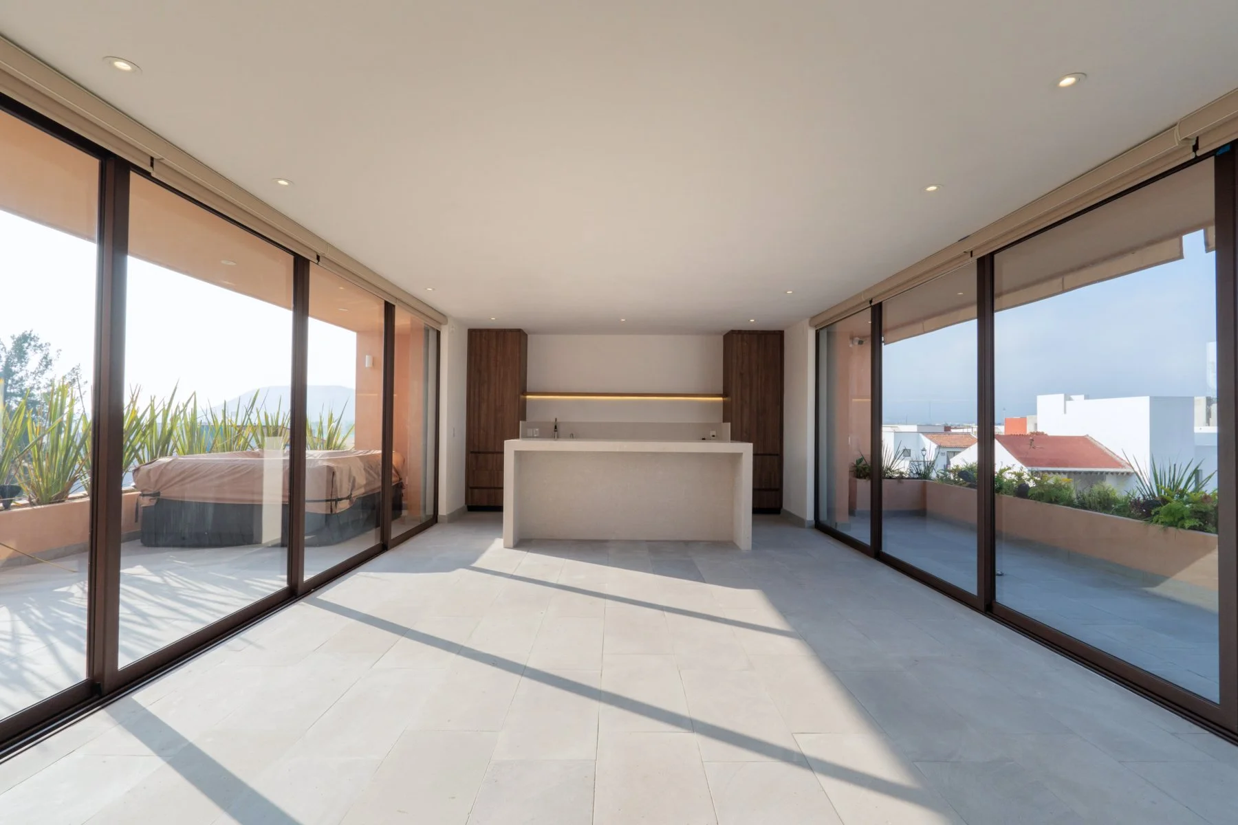 Modern indoor space with large glass sliding doors, an outdoor balcony with plants, a bathroom or kitchenette area with a minimalist design, light-colored flooring, and a view of neighboring rooftops and sky.