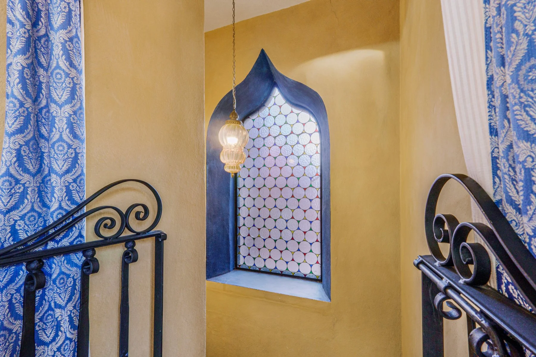 Decorative window with colorful geometric pattern, black wrought iron railing, yellow walls, blue patterned curtains, and a hanging glass lantern in a warmly lit room.