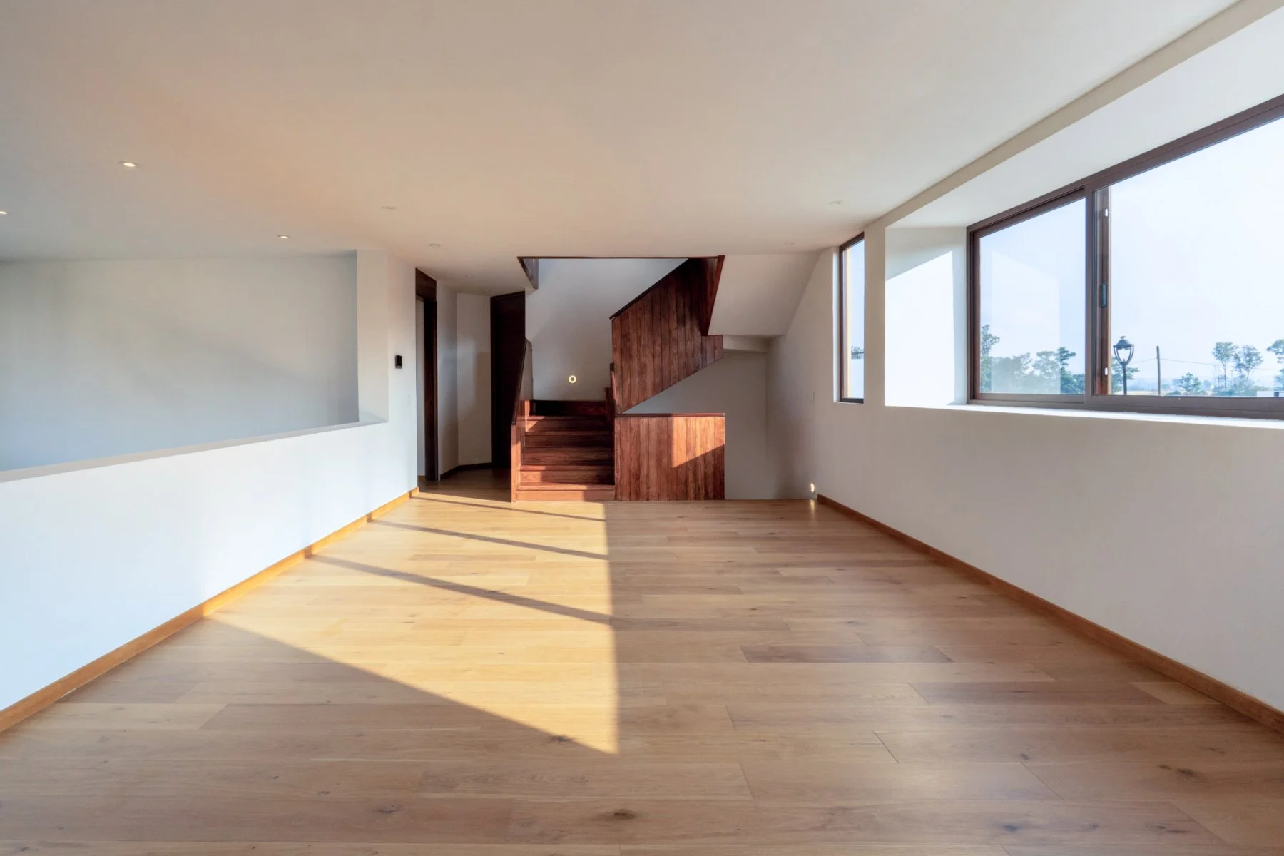 Empty living room with large windows, wooden flooring, and staircase with wooden steps and paneling.