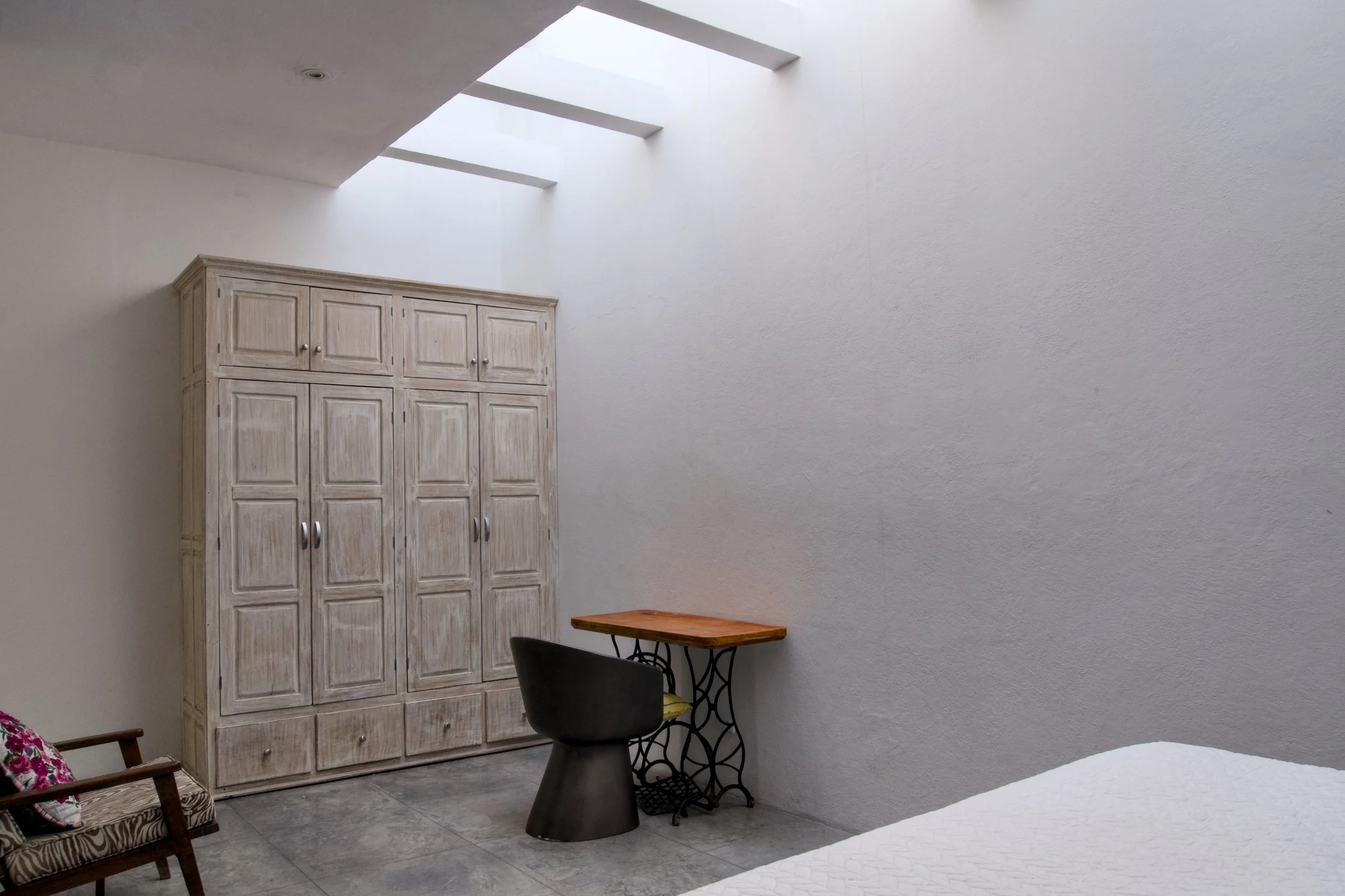 A bedroom corner with a large wooden wardrobe, a small wooden desk, a modern black chair, part of a bed with white bedding, a vintage armchair with floral cushions, and a ceiling with skylights letting in natural light.