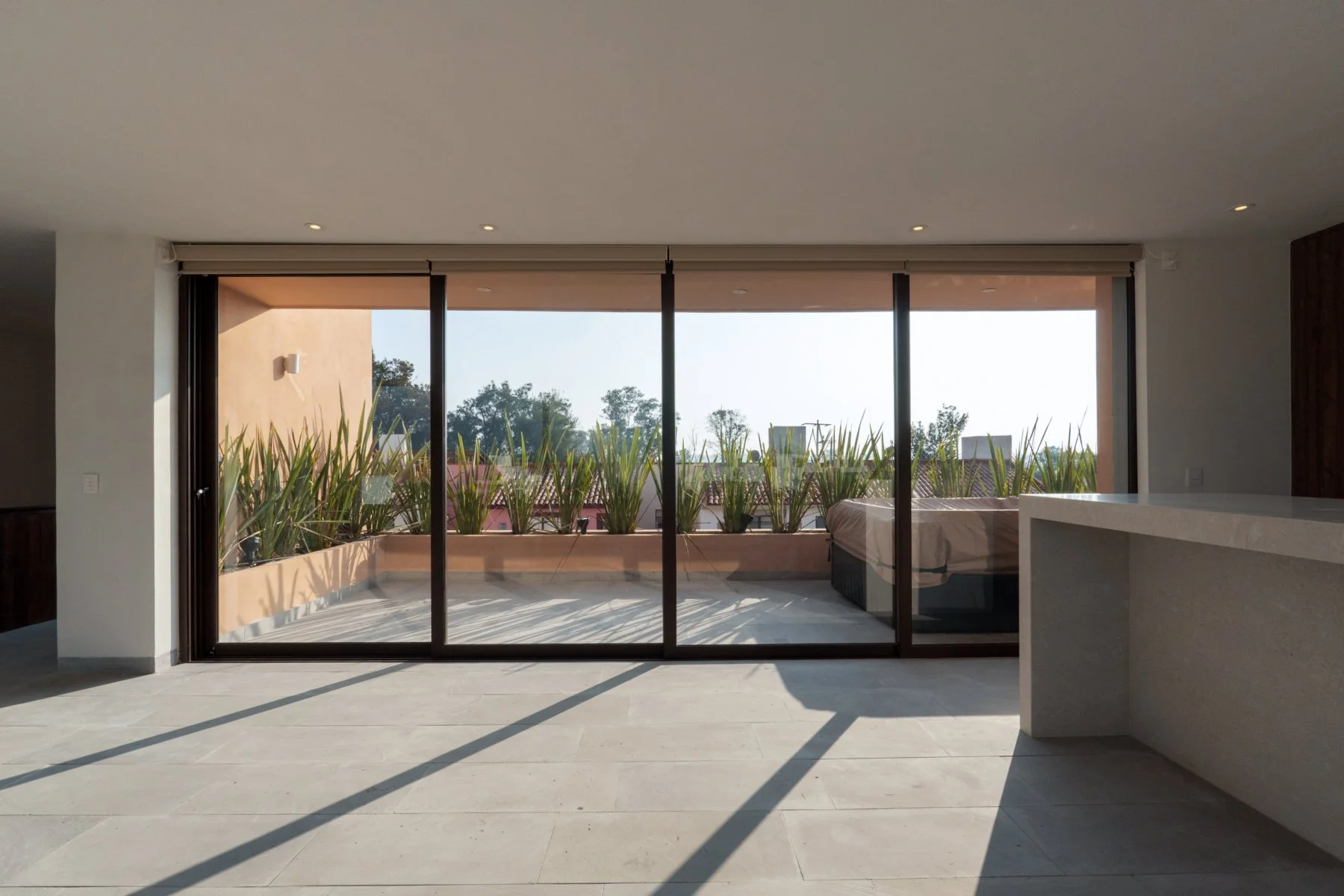Empty indoor living space with large glass sliding doors opening to a balcony with plants, sunlight casting shadows on the tiled floor.