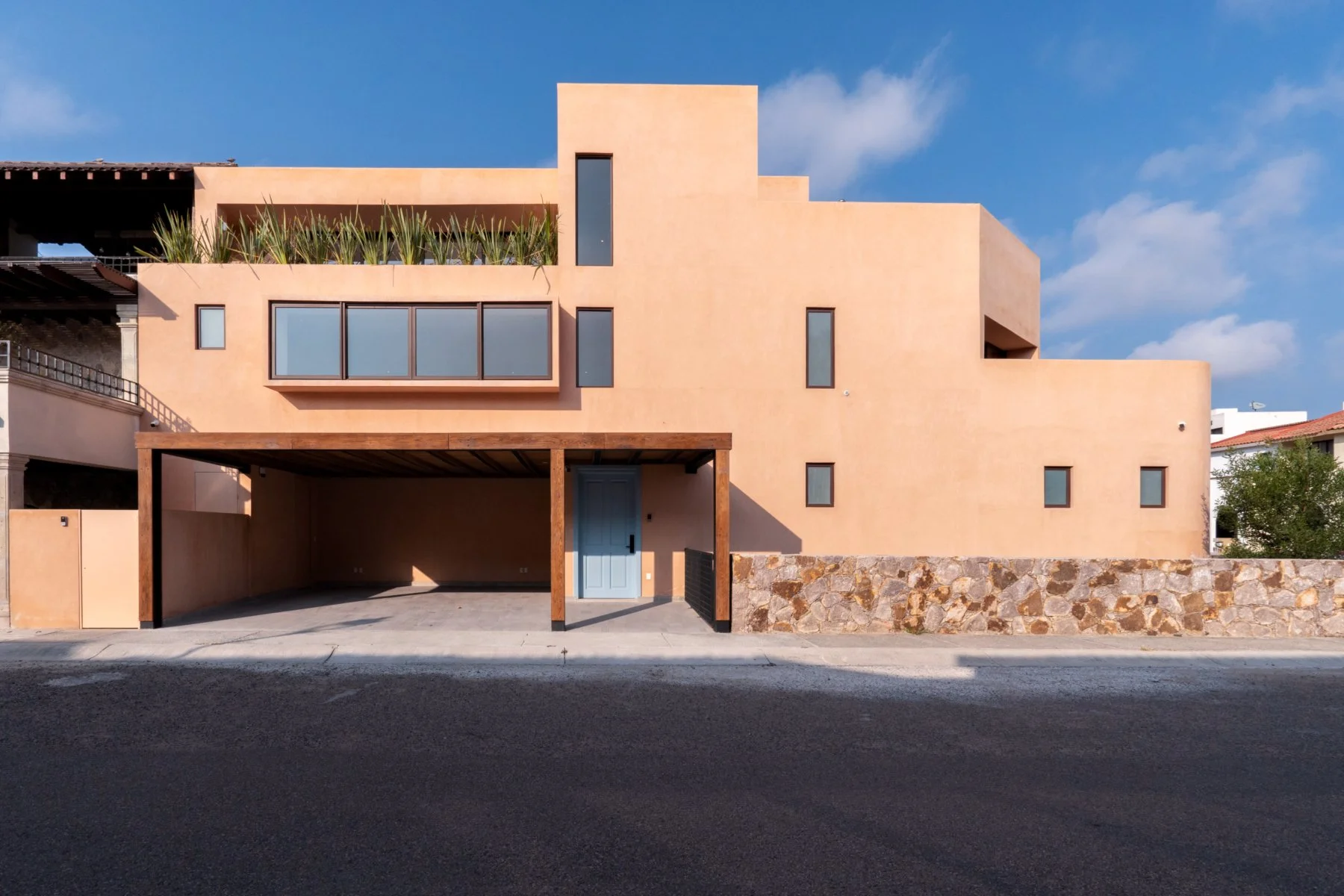 Modern peach-colored house with multiple varying-sized windows, a stone wall in front, and a blue gate, under a blue sky with some clouds.