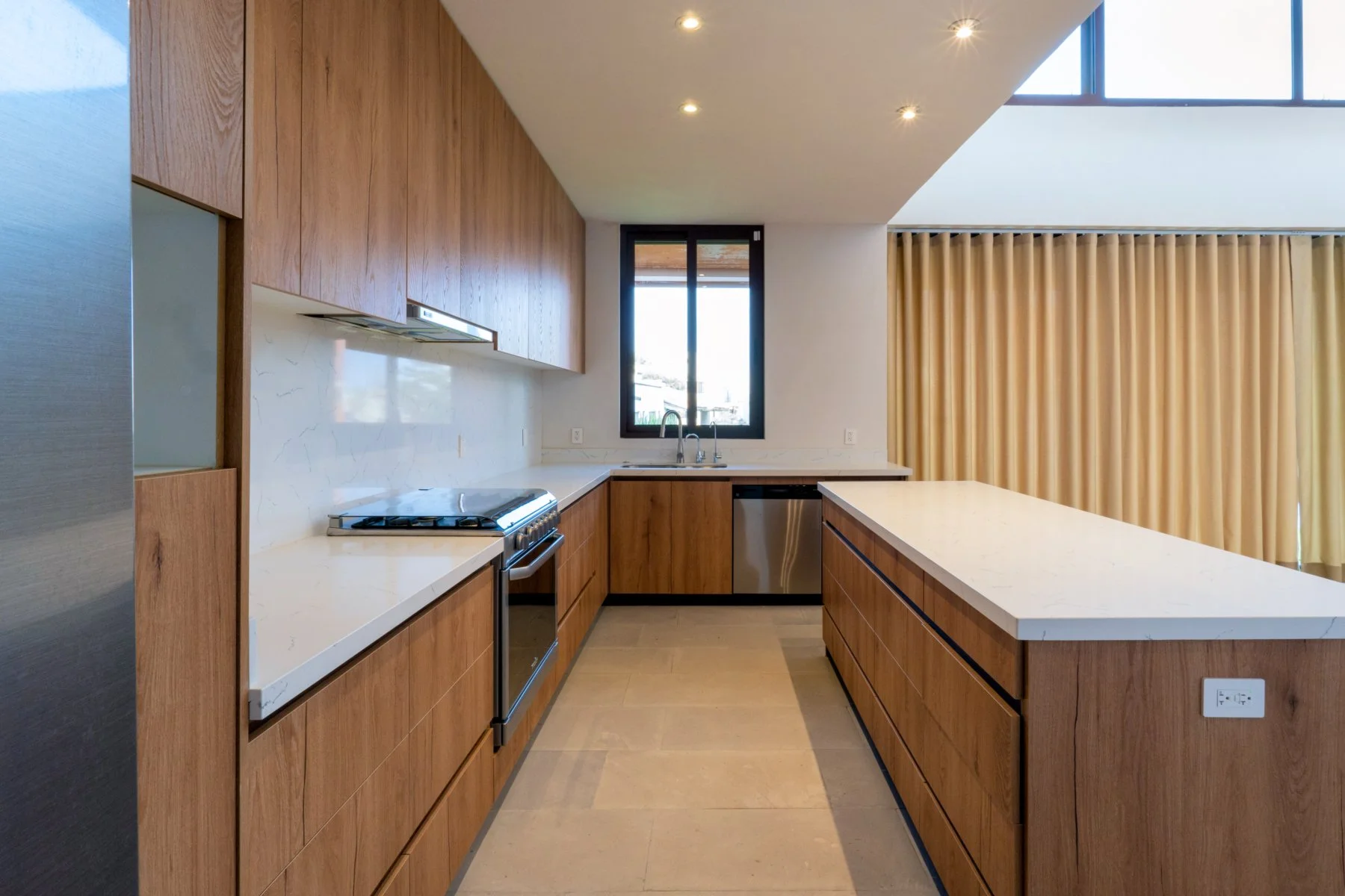 Modern kitchen with wooden cabinets, white countertops, stainless steel appliances, a window above the sink, and beige curtains in the background.