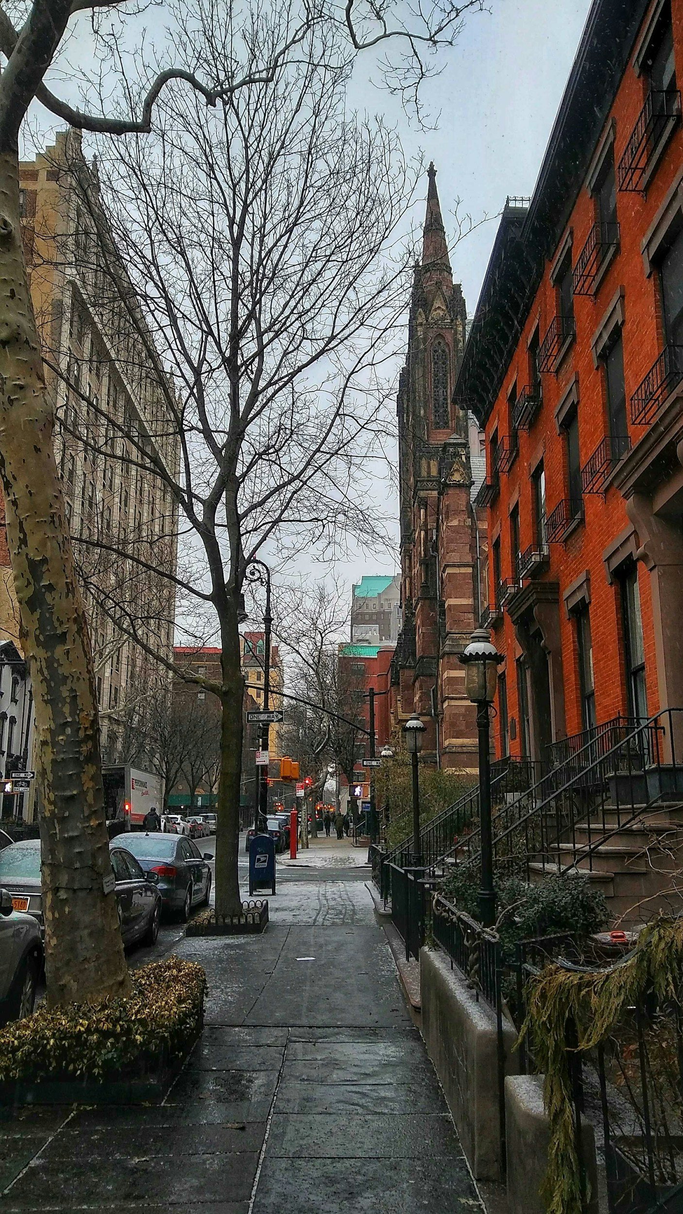 A city street scene with leafless trees, parked cars along the curb, and historic red brick buildings with black fire escapes. A tall church steeple rises in the background under an overcast sky.