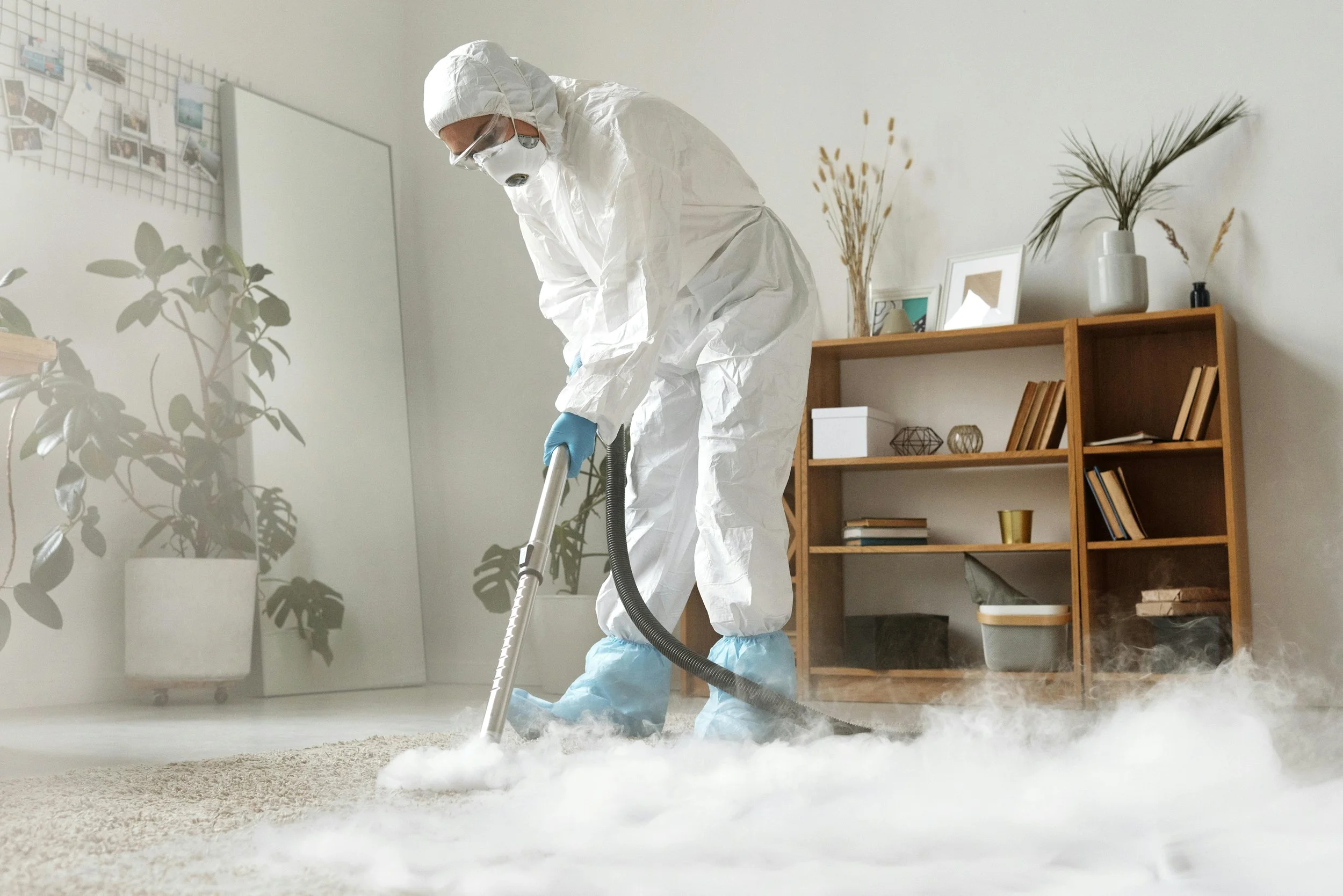 Person in protective gear using a fogger on a carpeted floor inside a room.