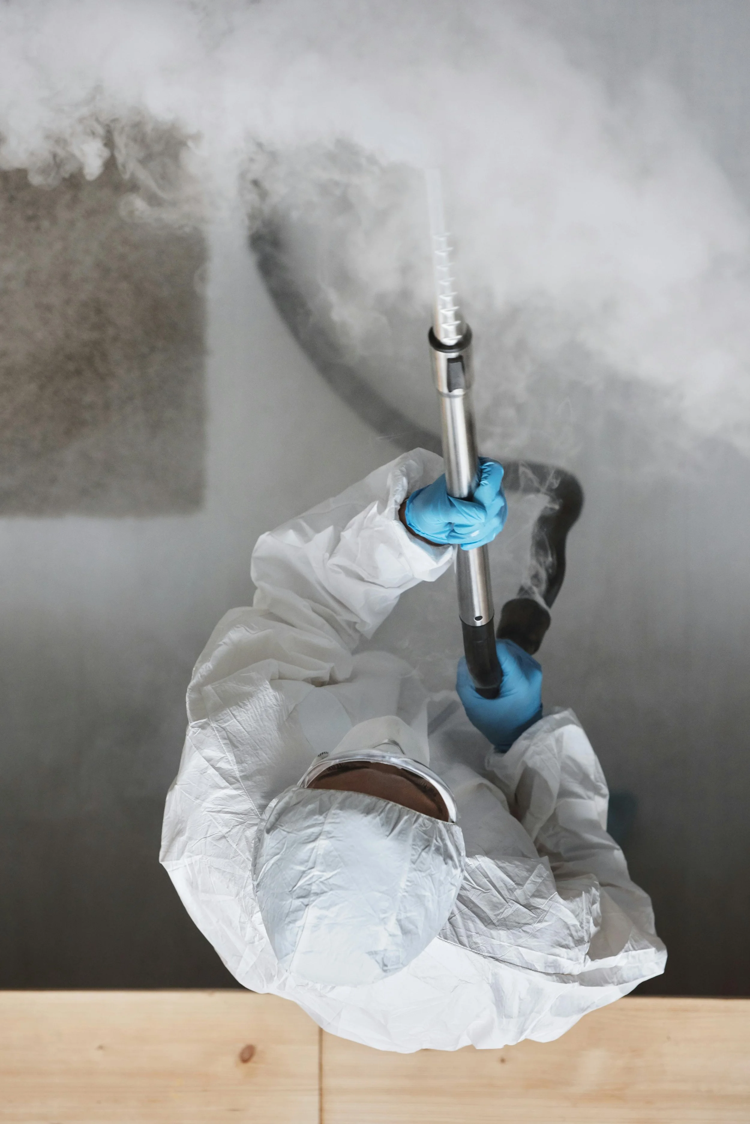 Person in protective suit and gloves holding a fogging or spraying device, emitting vapor or mist, in an indoor setting.
