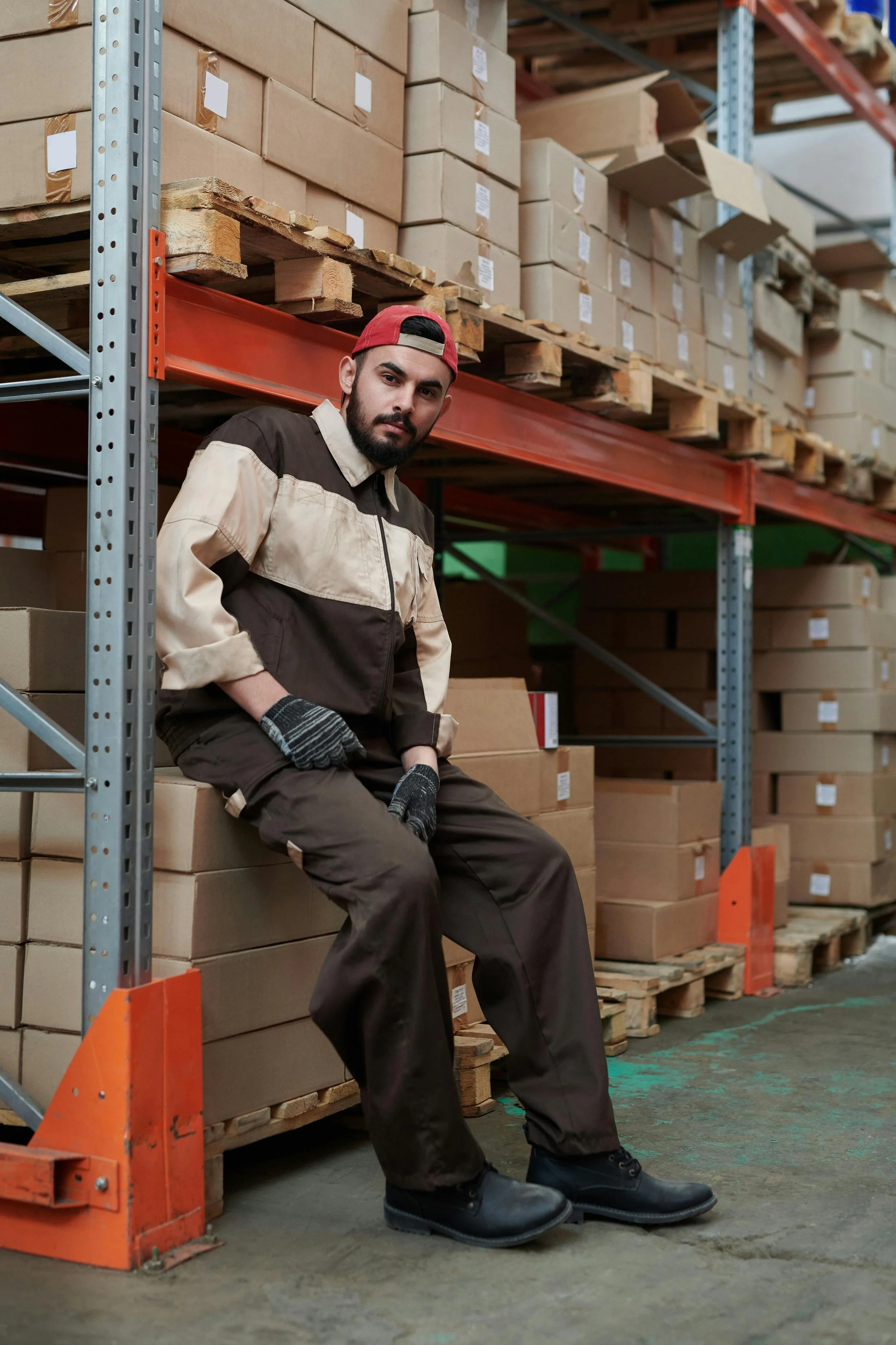 A man in work clothes sitting on a stack of cardboard boxes in a warehouse, surrounded by metal shelving filled with stored boxes.