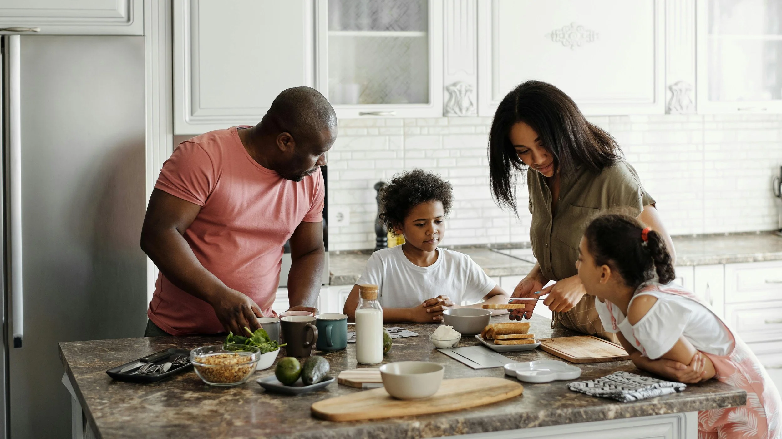 A family in the kitchen preparing breakfast together. A man, woman, and two young girls are gathered around a kitchen island with various food items and dishes.