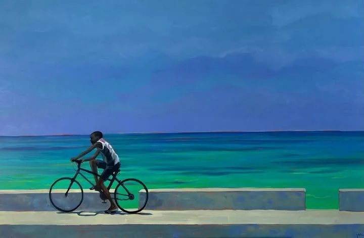 A man riding a bicycle on a coastal road with a large body of water and a cloudy sky in the background.