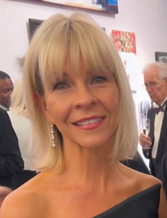 A smiling middle-aged woman with blonde hair and earrings at a social gathering.