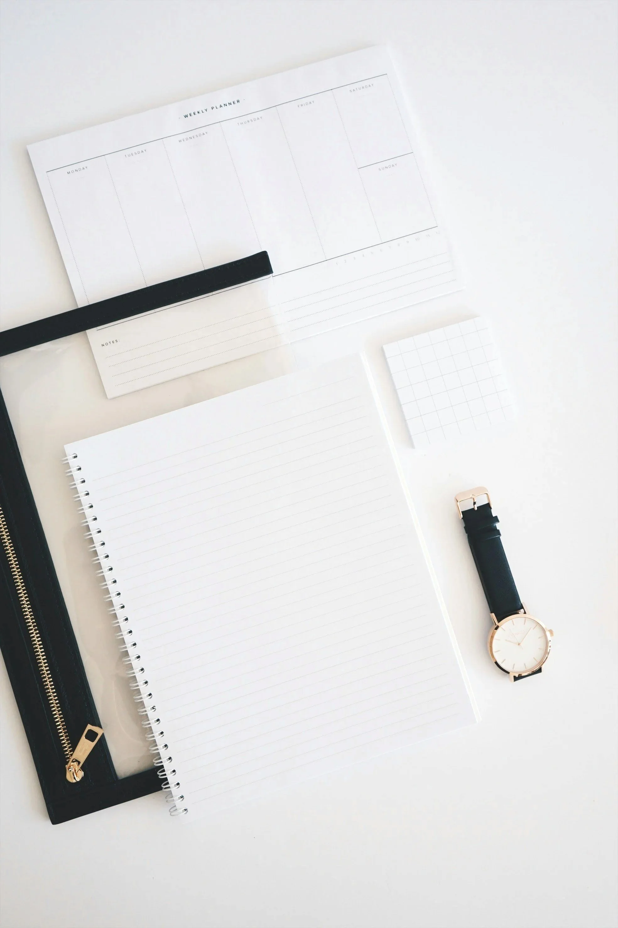 Flat lay of a minimalist planner, notebook, small notepad, watch, black zipper pouch, and pen on white surface.