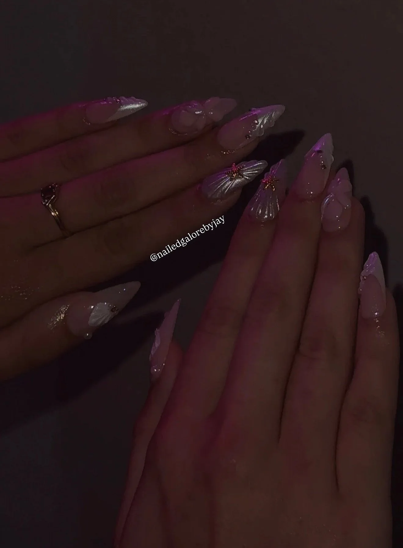 Close-up of two hands with decorated, glossy, pinkish-white nails, some with 3D flower designs and small embellishments, set against a dark background.