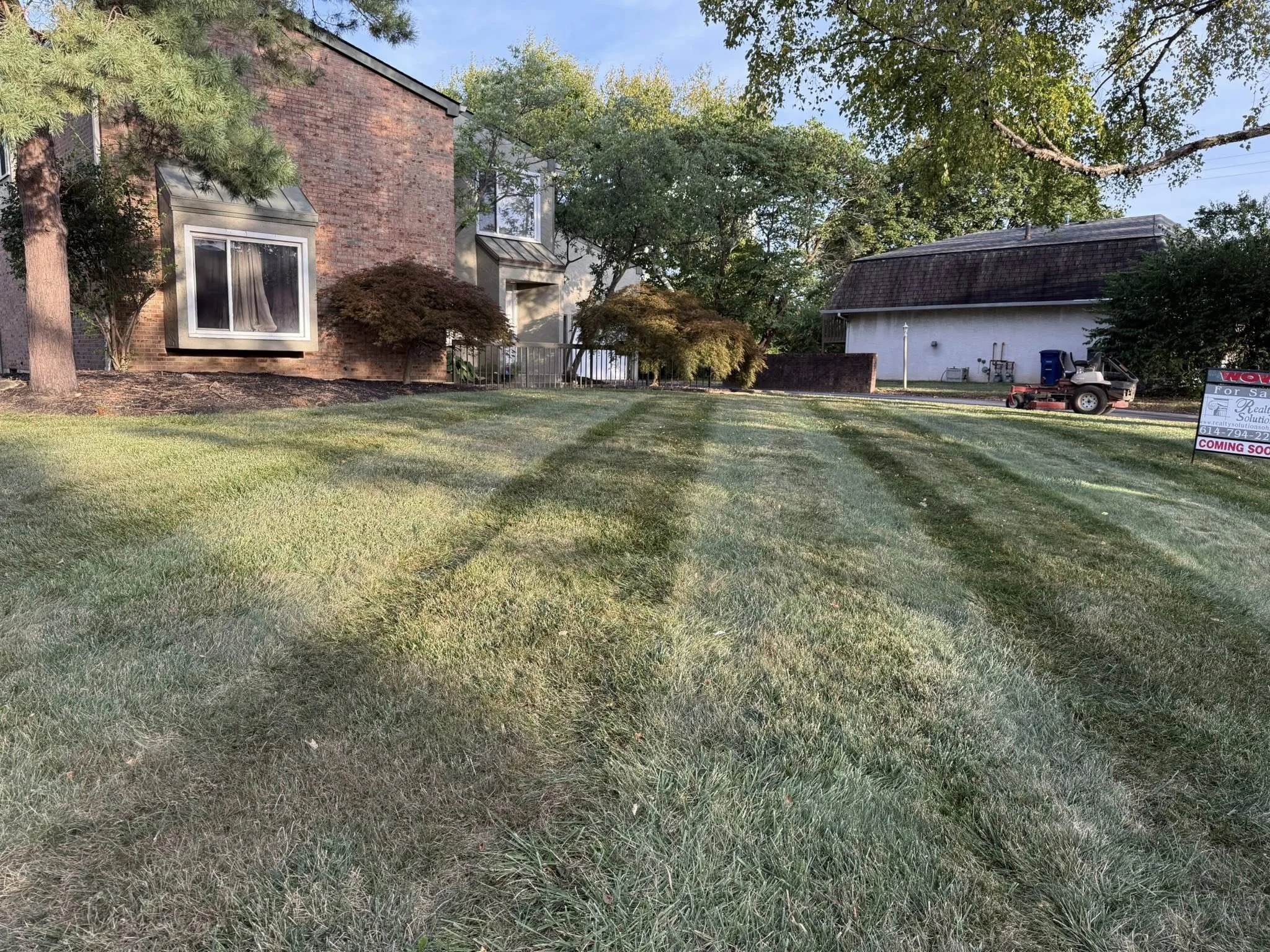 Well-maintained green lawn with tire tracks, a brick building with bay windows, trees, and a white building with a roof in the background, a lawnmower, and a 'Coming Soon' real estate sign.