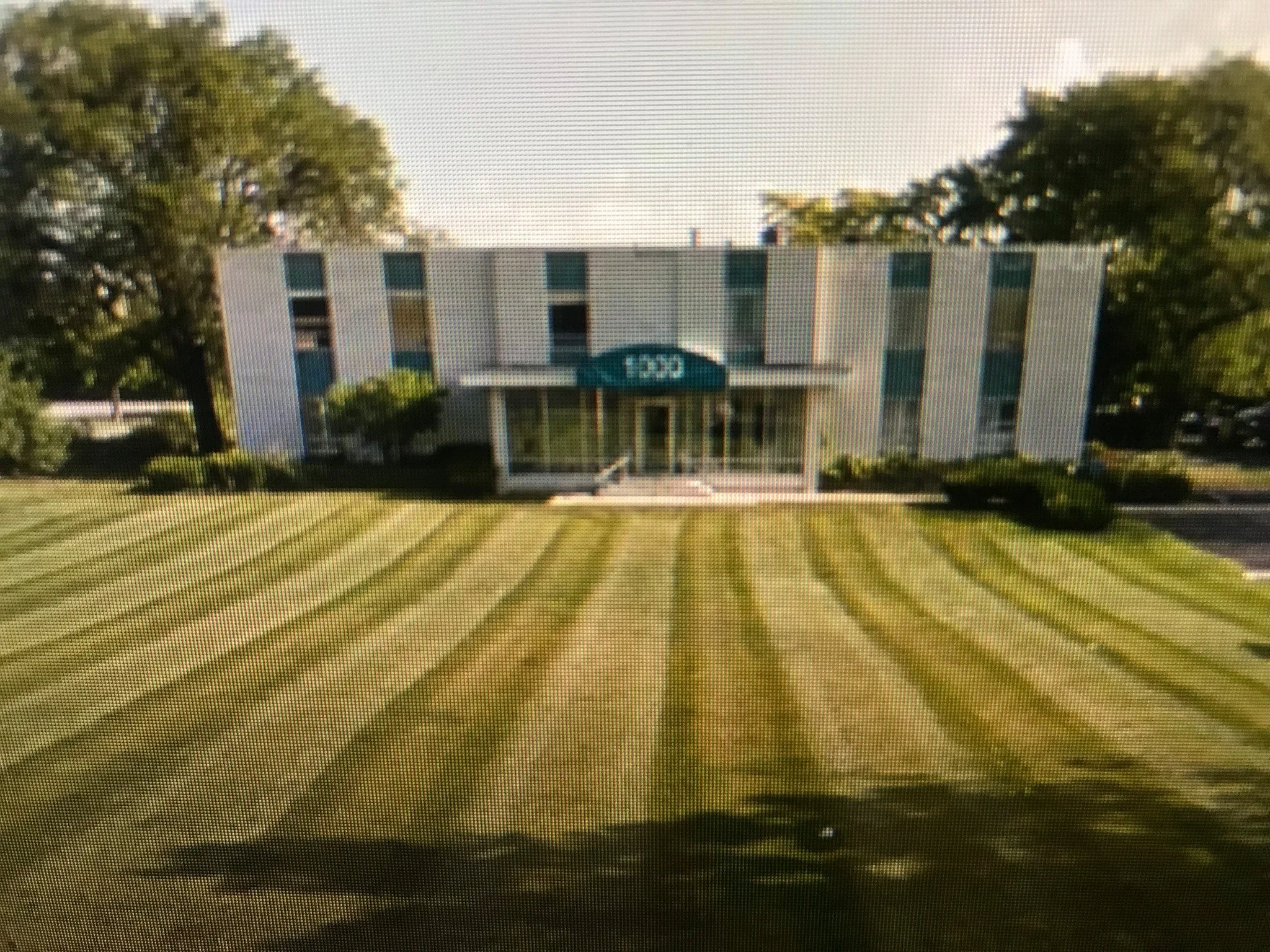 A multi-story office building with a blue awning and large windows, surrounded by a well-maintained lawn with striped grass patterns.