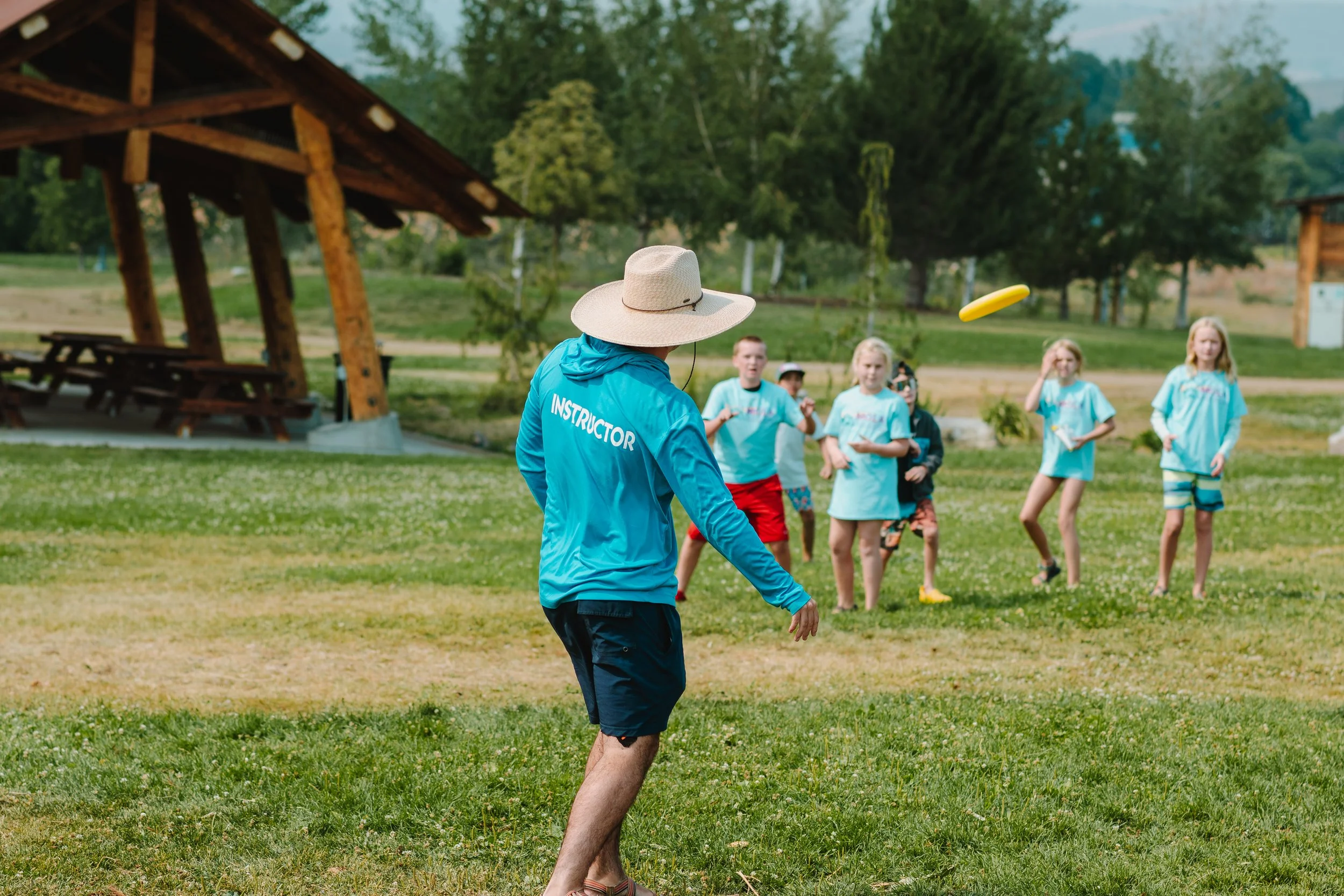 Instructor wearing a blue sun hoodies and wide-brimmed hat leading children in a game of frisbee outdoors in a park.