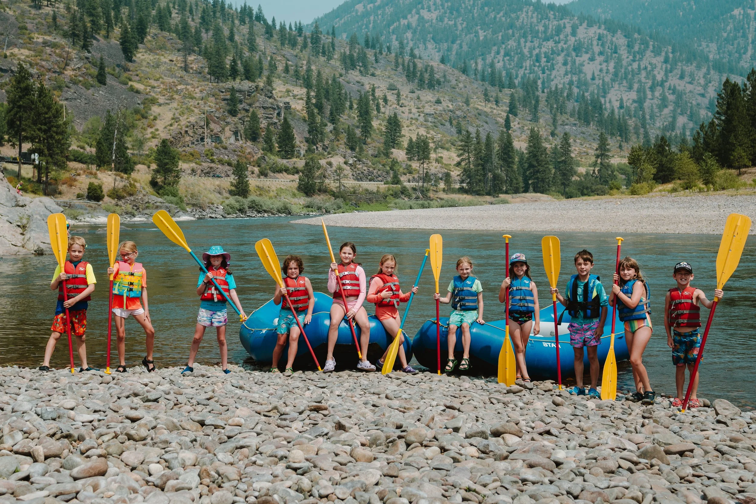 Group of children in life jackets holding paddles standing on rocky shoreline next to blue inflatable raft with mountains and trees in the background.