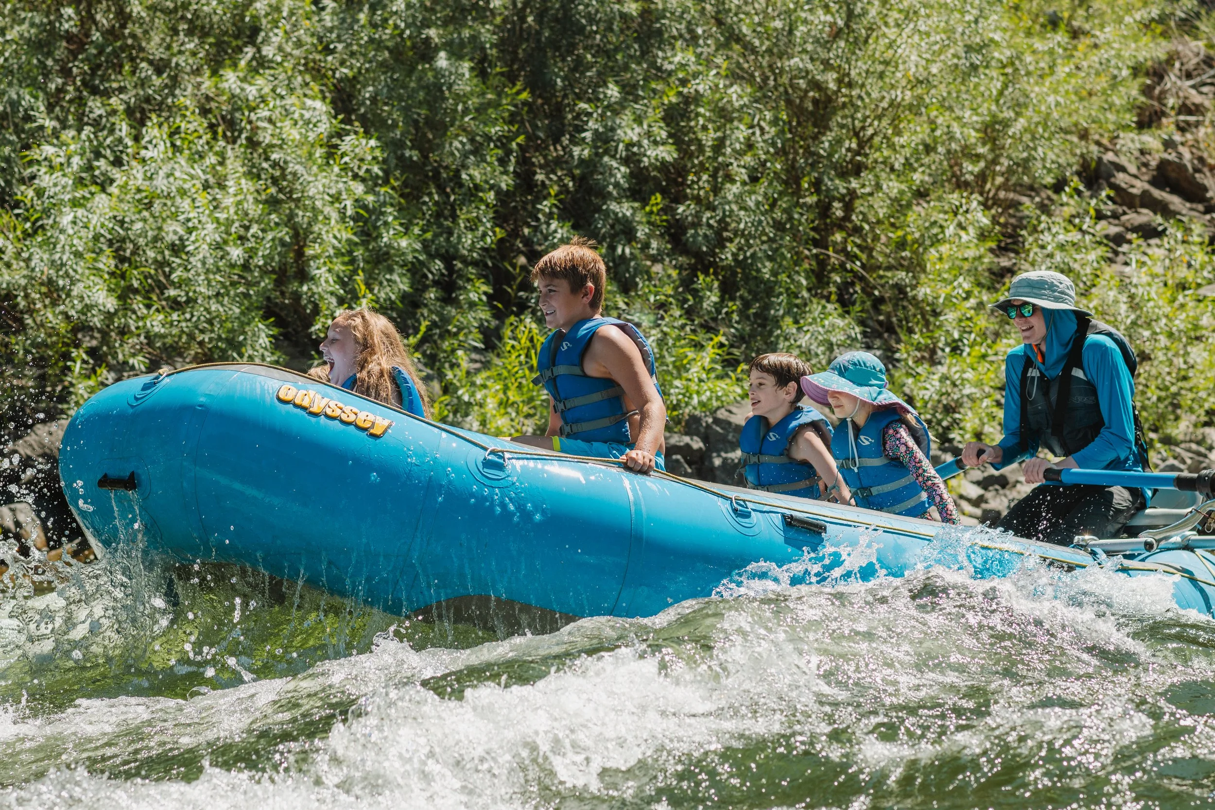 Group of children and a woman whitewater rafting on a river surrounded by green trees.