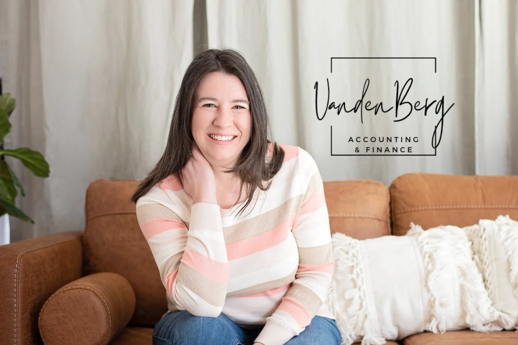 A woman with long dark hair, smiling, wearing a striped beige, pink, and white sweater, sitting on a brown couch with decorative pillows in a cozy living room. There is a plant on the left and a sign that says "VandenBerg ACCOUNTING & FINANCE" on the right.