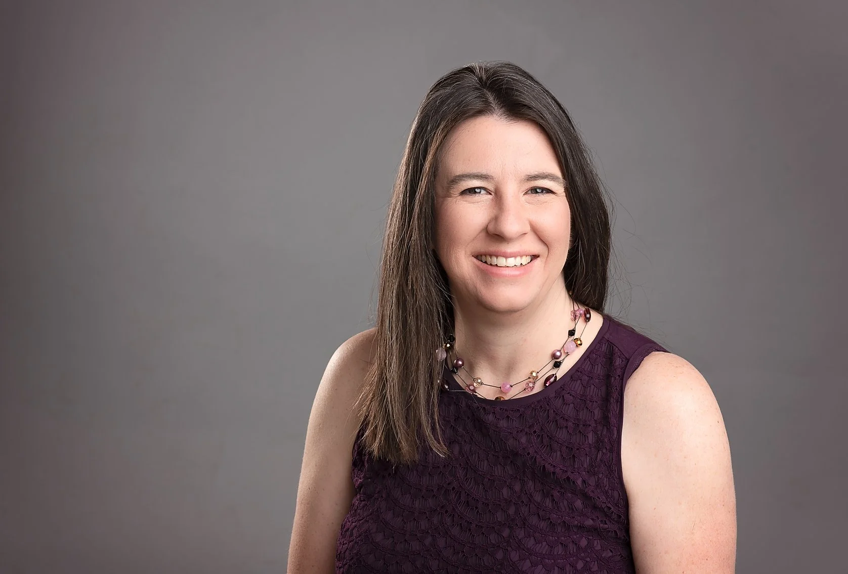 A woman with shoulder-length dark hair smiling against a gray background, wearing a purple sleeveless dress and a multi-strand necklace with beads.